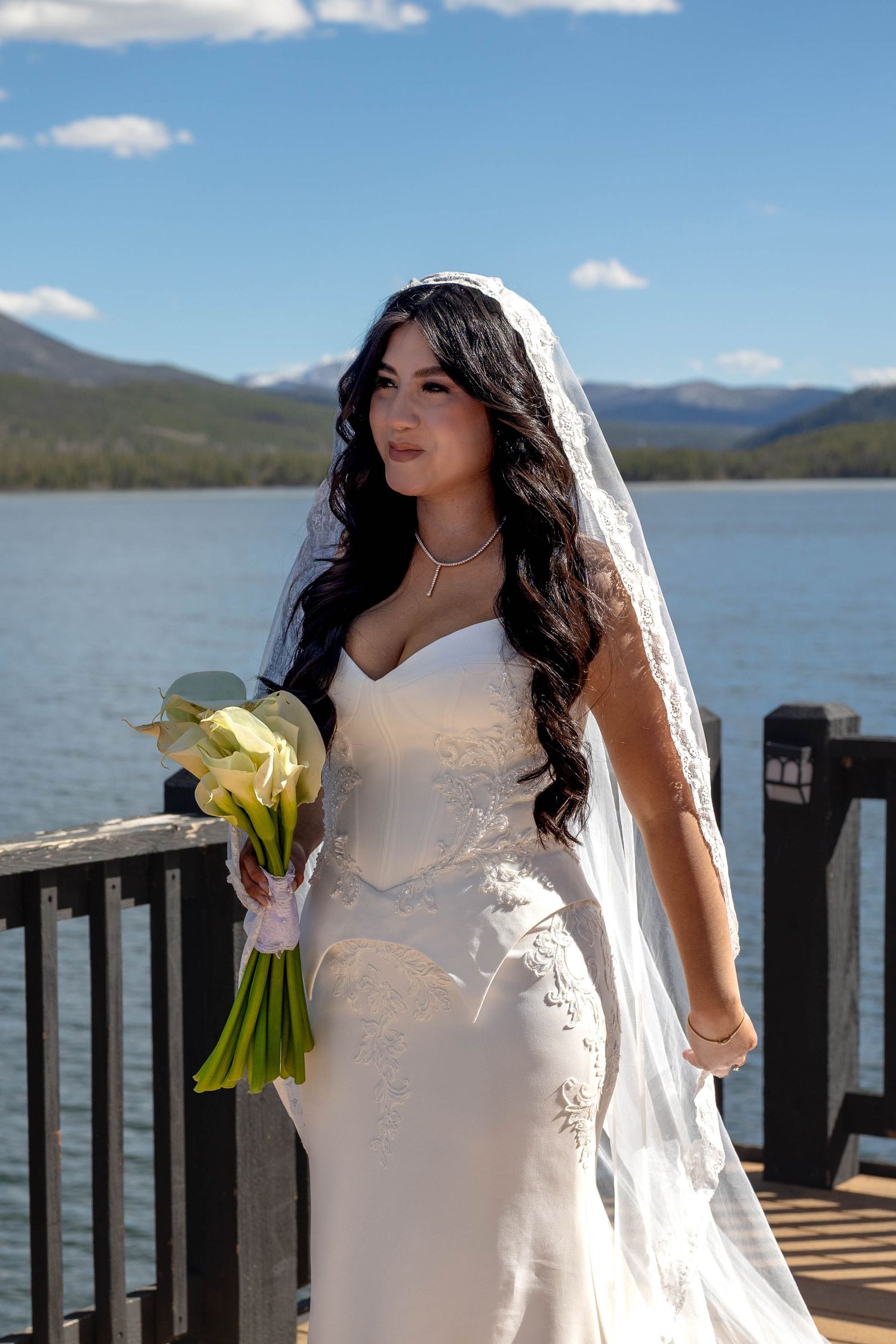 Bride in white lace gown and veil holds calla lily bouquet on lakeside deck with mountain backdrop.