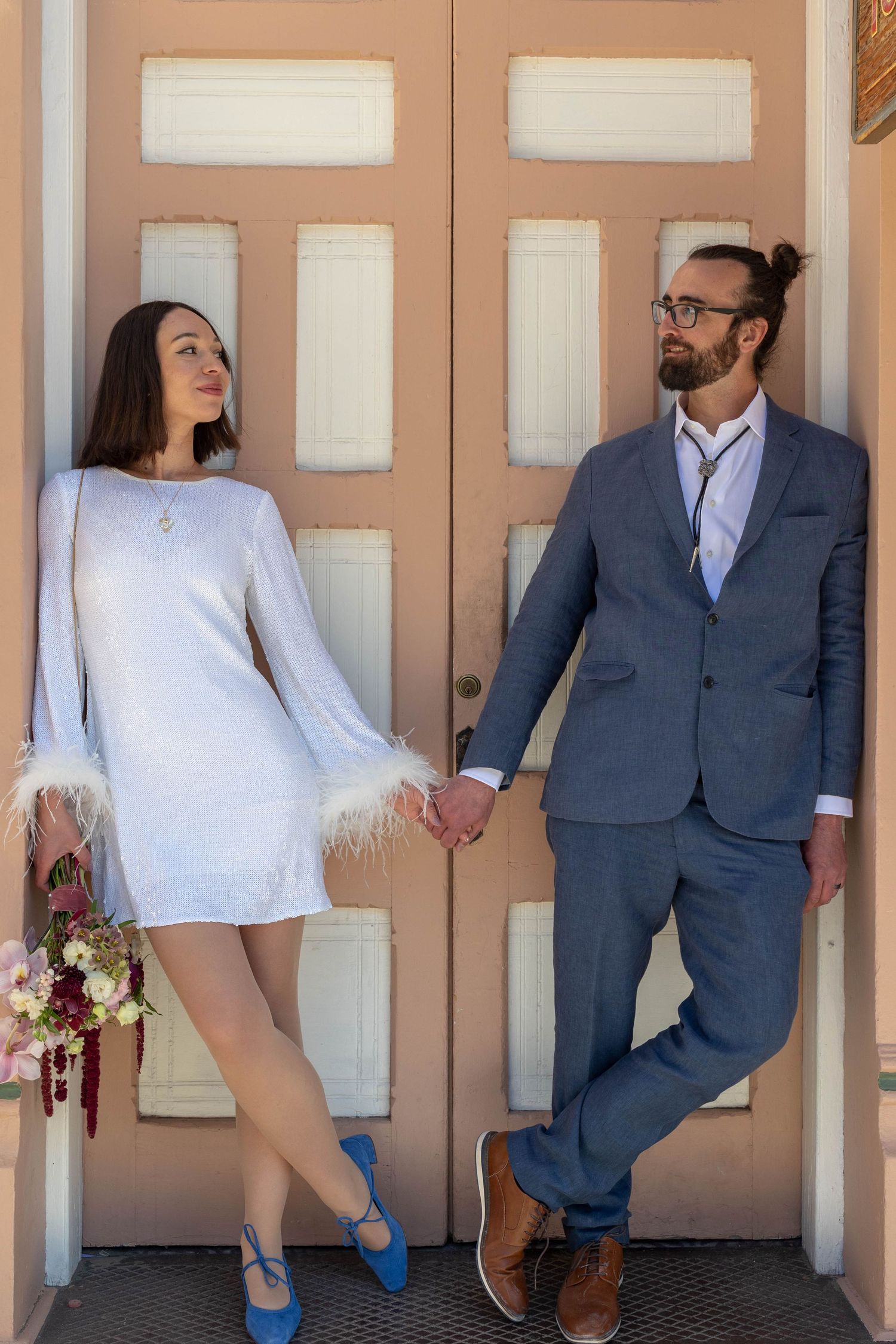 Bride in white feather-trim mini dress with blue heels holds groom's hand in blue suit before pink double doors.