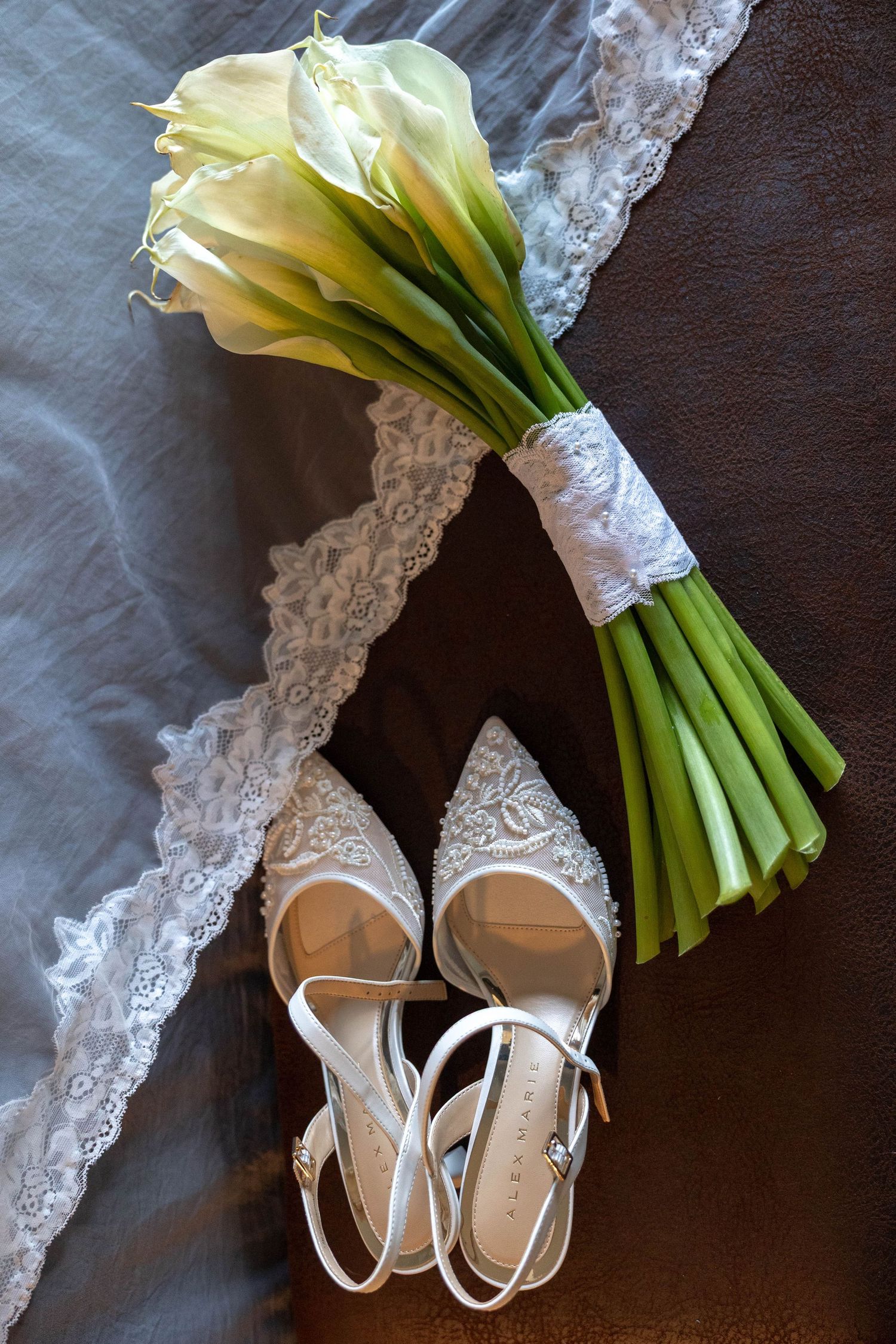 White lace bridal shoes and calla lily bouquet with lace veil on dark background for wedding day.
