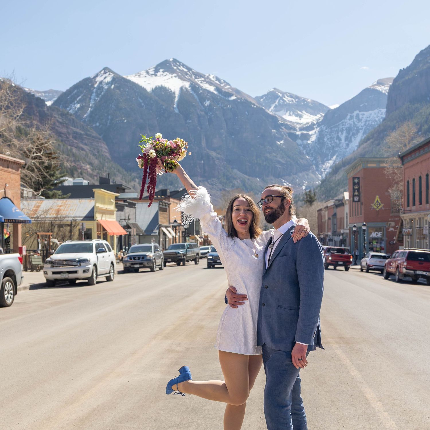 Joyful newlyweds celebrate on a mountain town street, bride raising bouquet, snow-capped peaks in background.