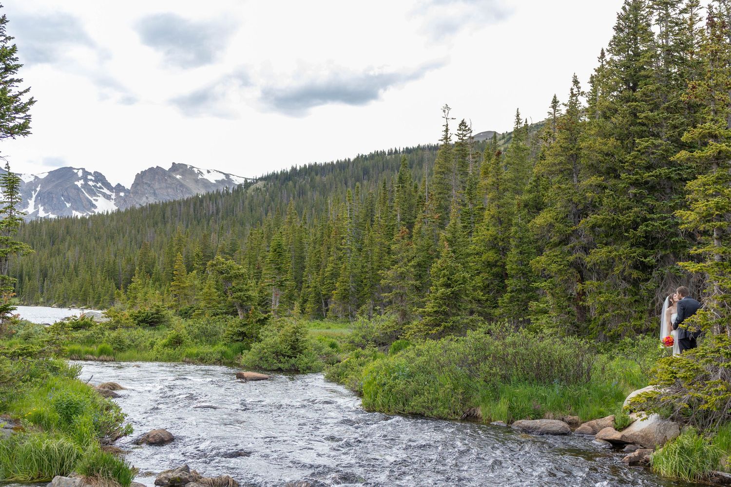 Hiker standing beside a rushing mountain stream surrounded by evergreen forest with snow-capped peaks in the background.
