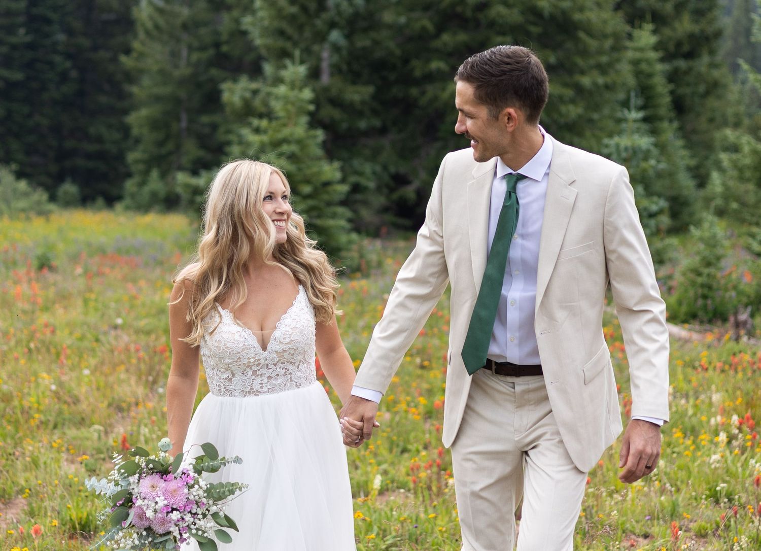 Couple walking hand in hand through vibrant mountain wildflower meadow in summer wedding attire.
