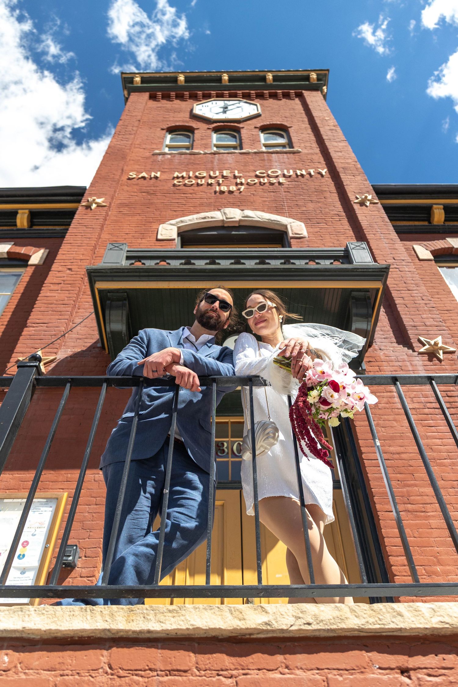 Bride and groom pose on balcony of historic San Miguel County Courthouse brick building under blue sky.
