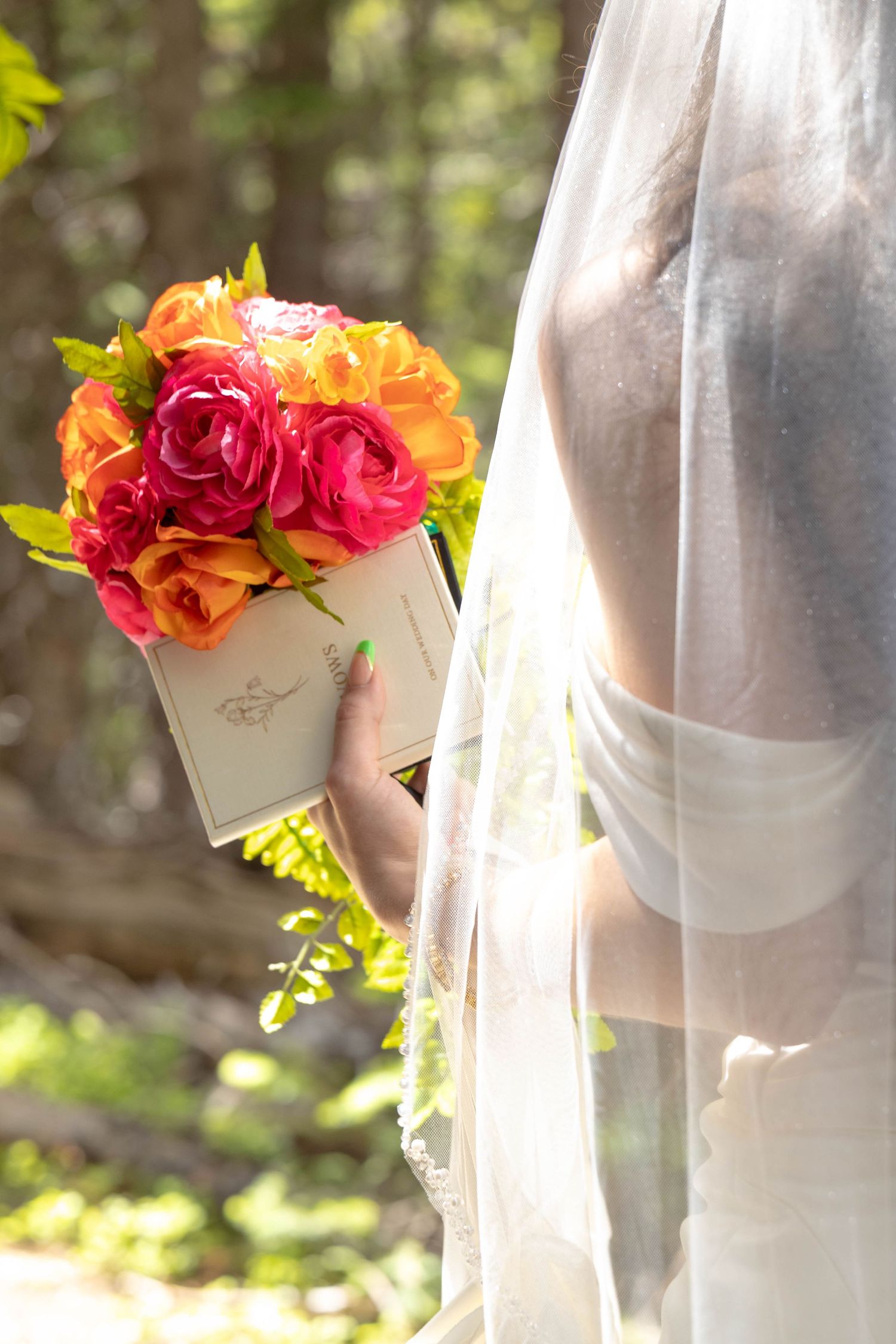 Bride in white wedding dress and veil holding vibrant pink and orange rose bouquet outdoors in natural forest setting.