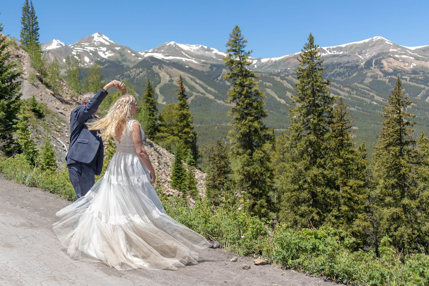 Bride and groom walking hand in hand with stunning Rocky Mountain peaks and evergreen trees in the background.