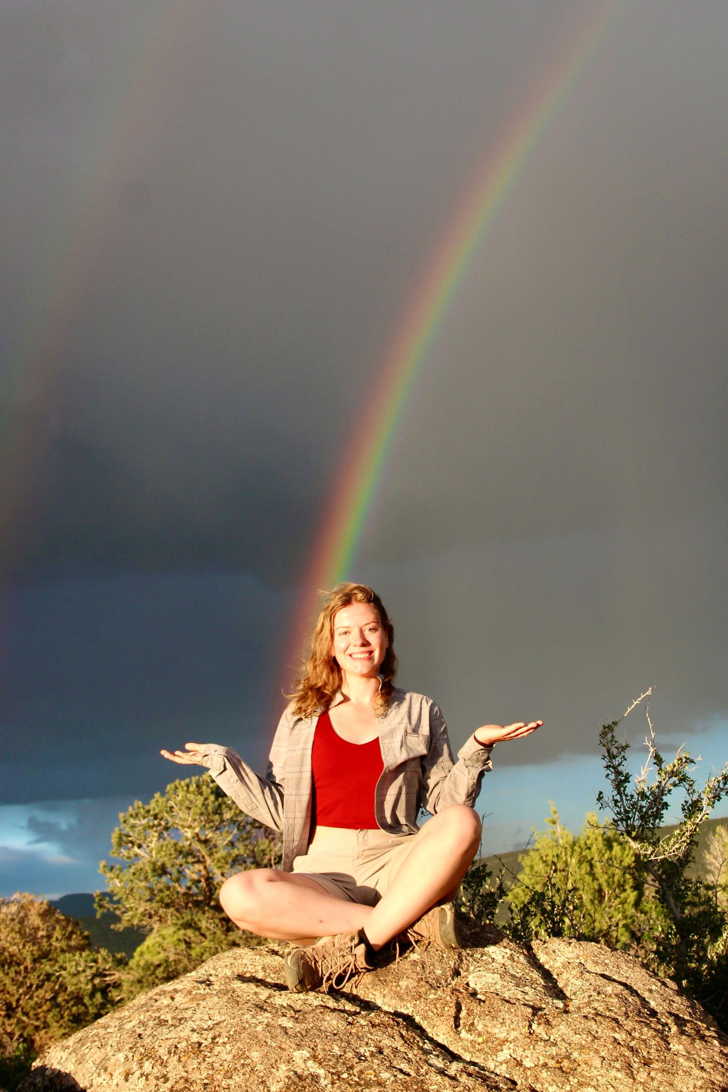 A smiling woman sits cross-legged on a rock with arms raised, framed by a stunning double rainbow in a dramatic sky.