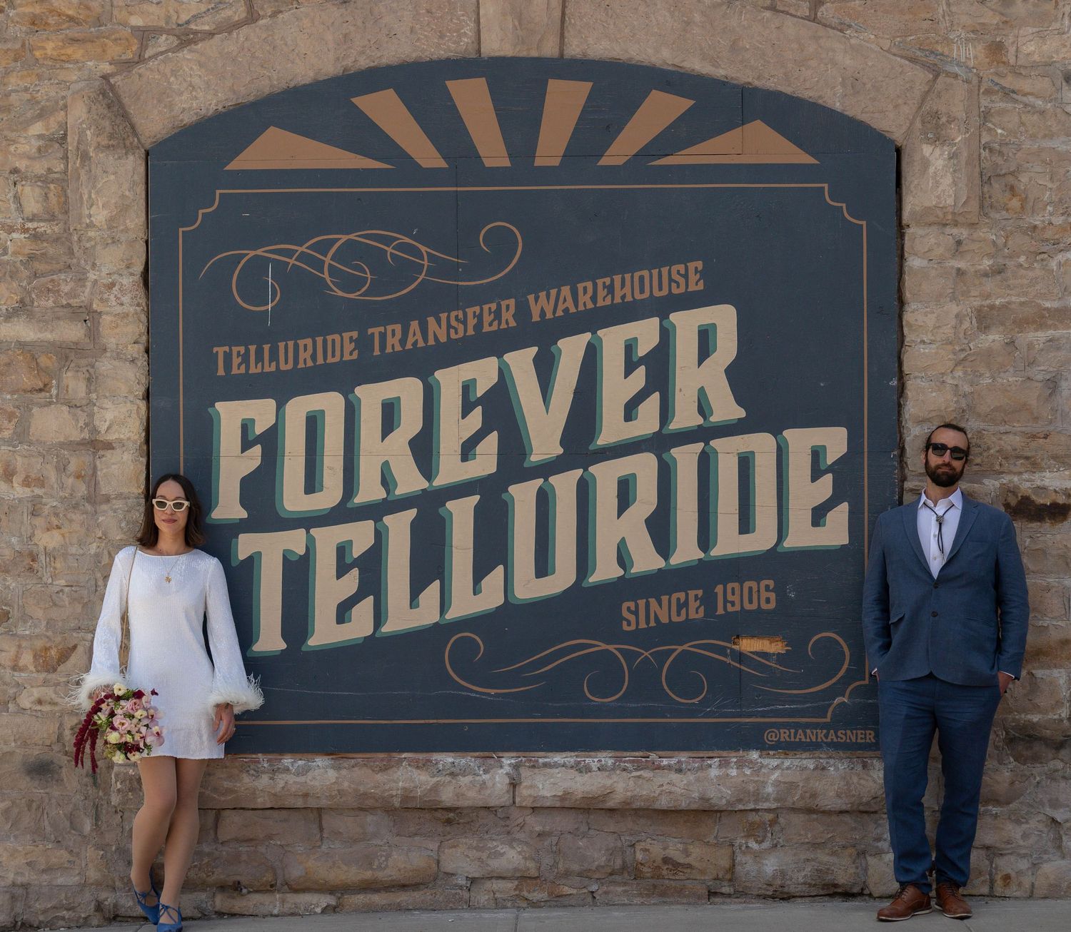 Couple posing in front of Forever Telluride Transfer Warehouse sign on stone wall in Telluride, Colorado.