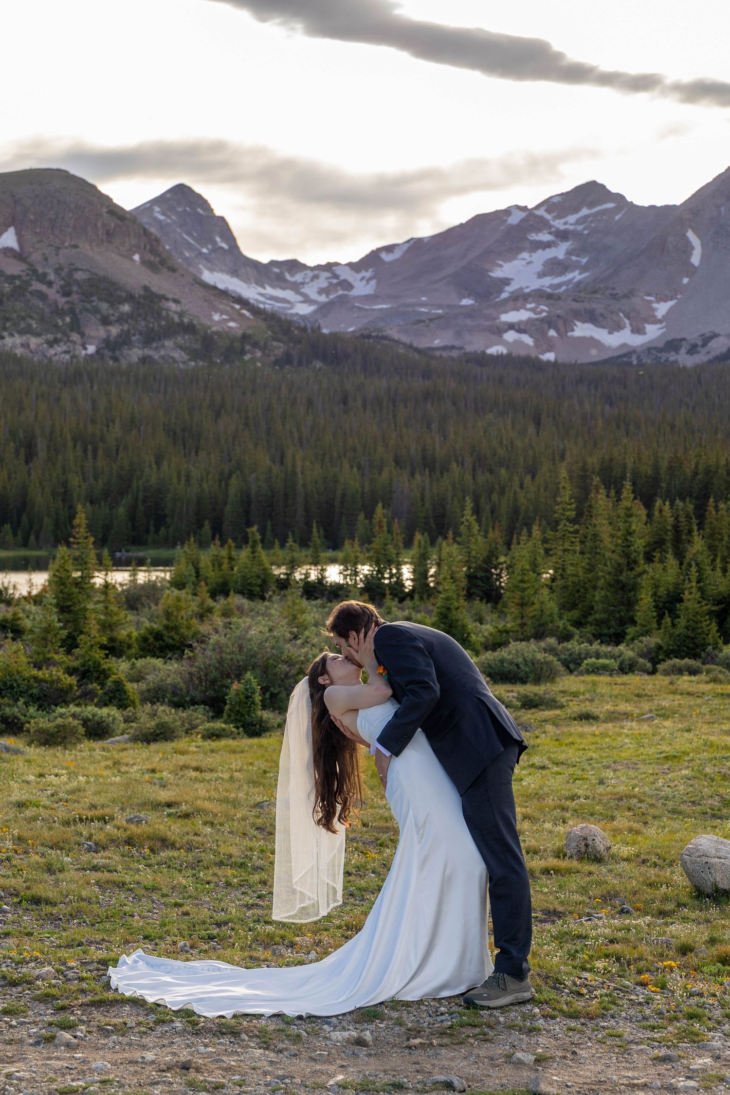 Bride and groom share a romantic dip kiss with stunning Rocky Mountain peaks and pine forest backdrop at sunset.