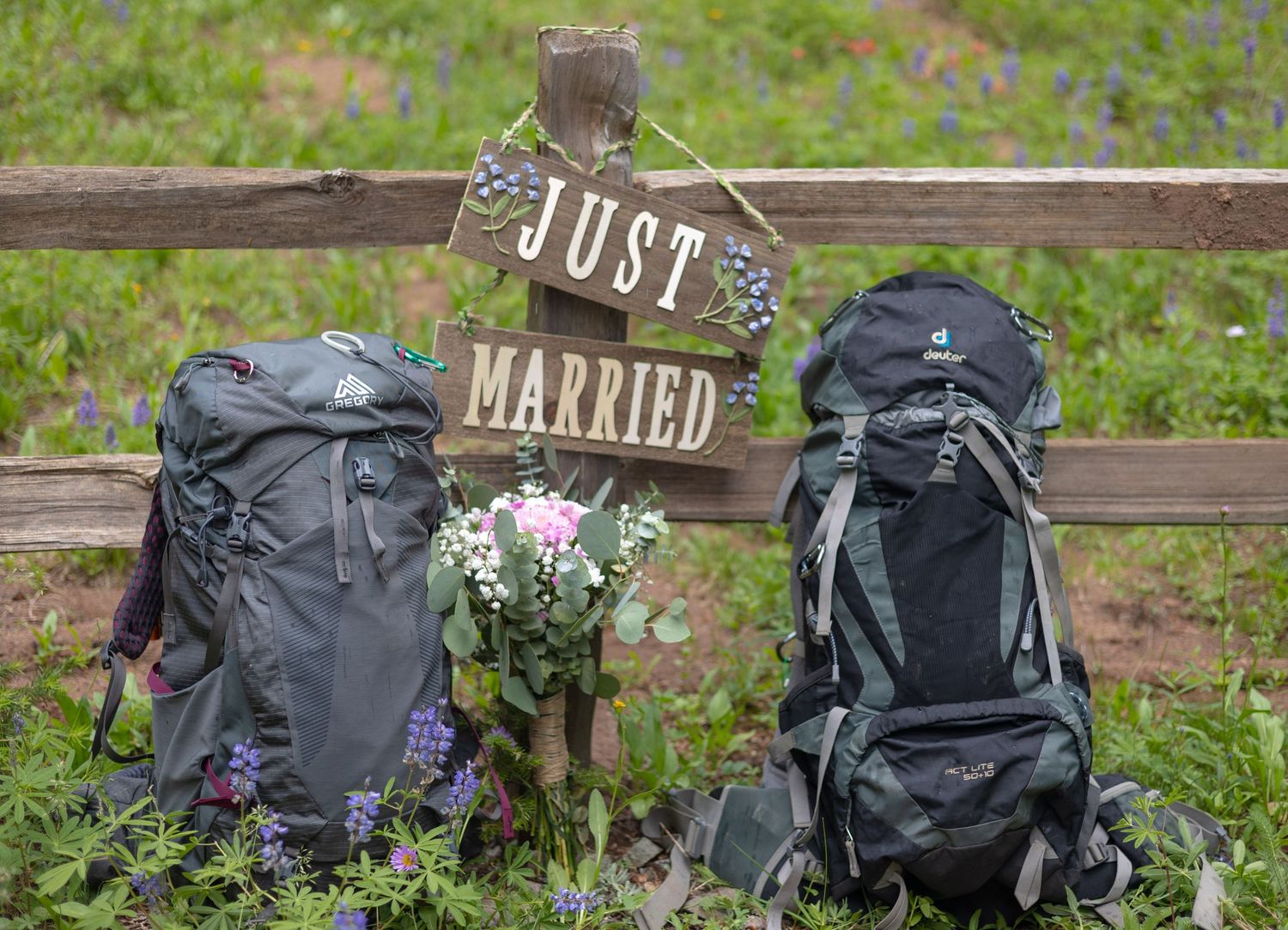 Two hiking backpacks on a wooden fence with a 'Just Married' sign and small floral bouquet between them.