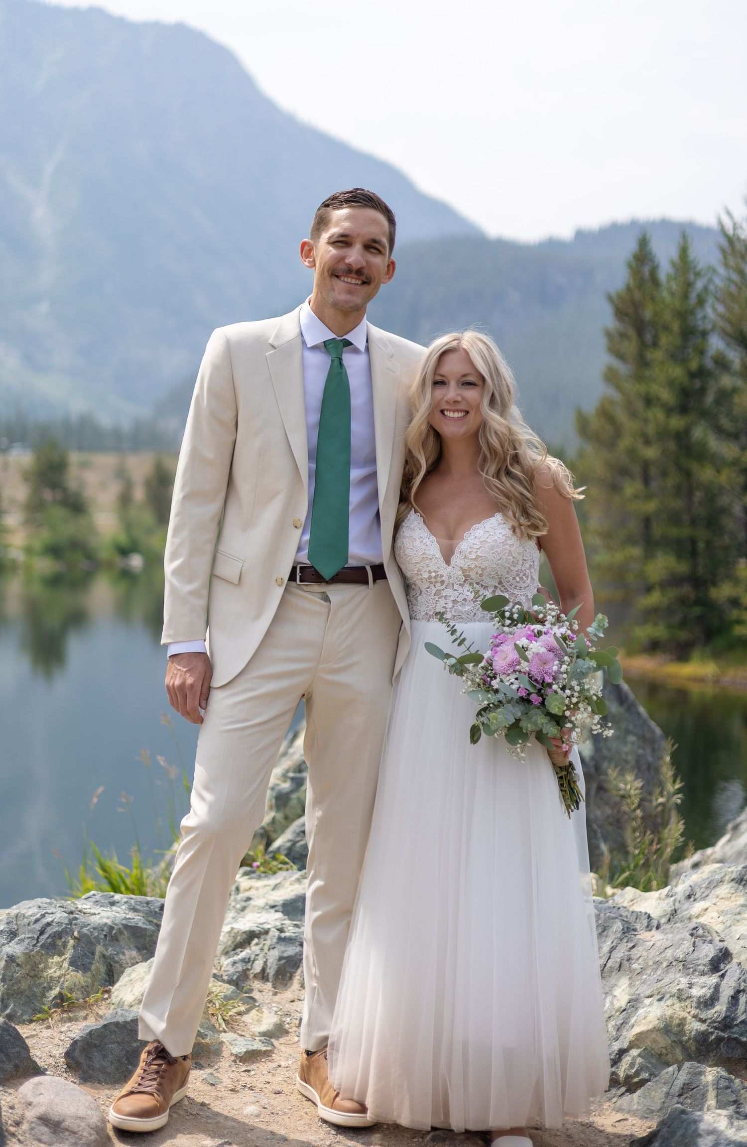 Wedding couple stands on rocky overlook with scenic mountain vista and lake behind them.