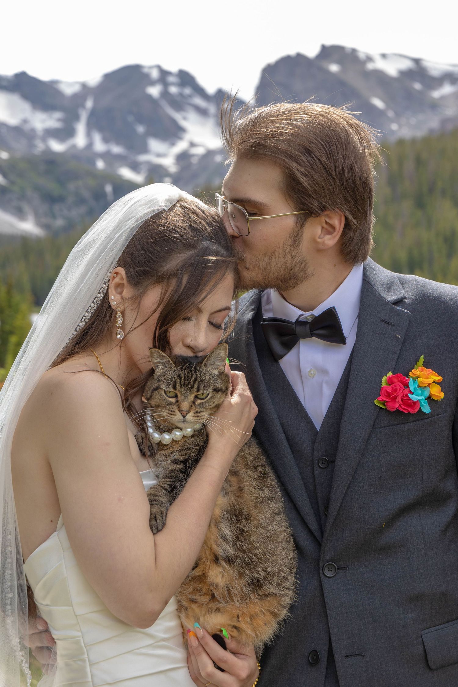 Bride and groom holding a tabby cat on their wedding day with snowy mountains in the background.