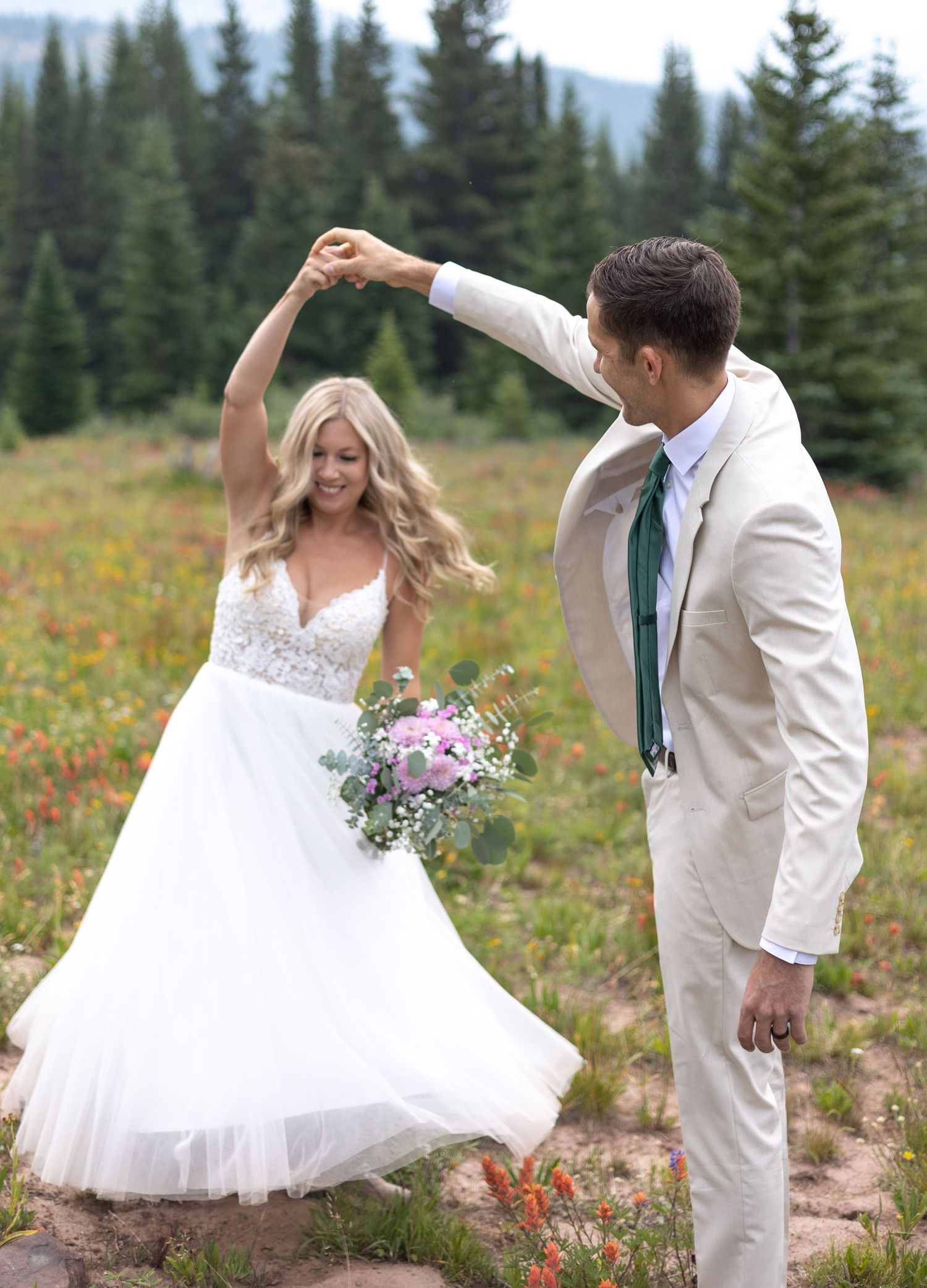 A couple in wedding attire dances and twirls together in a mountain meadow filled with wildflowers and evergreen trees.