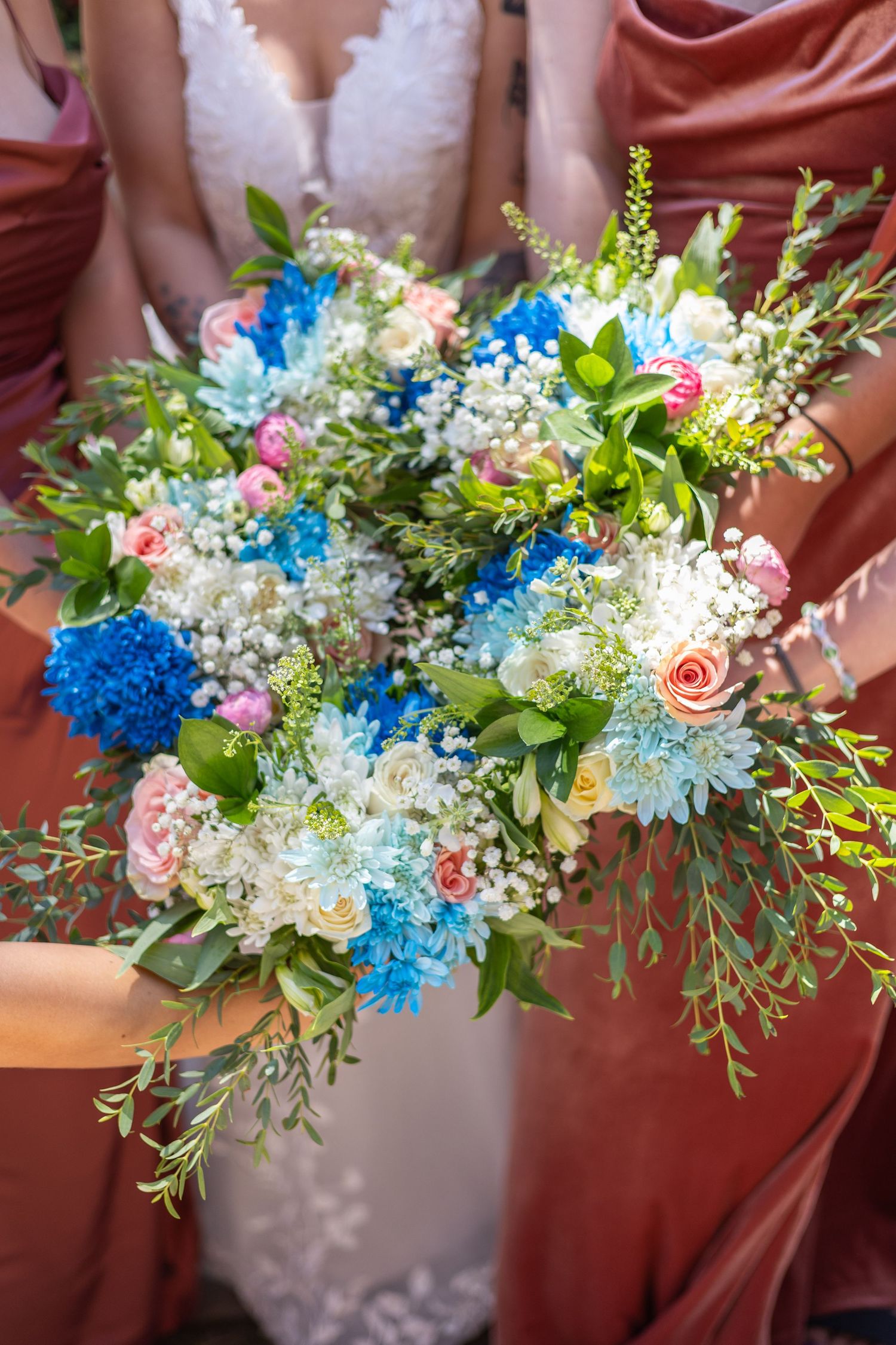 A vibrant wedding bouquet featuring blue carnations, pink roses, white baby's breath and green foliage against burgundy fabric.