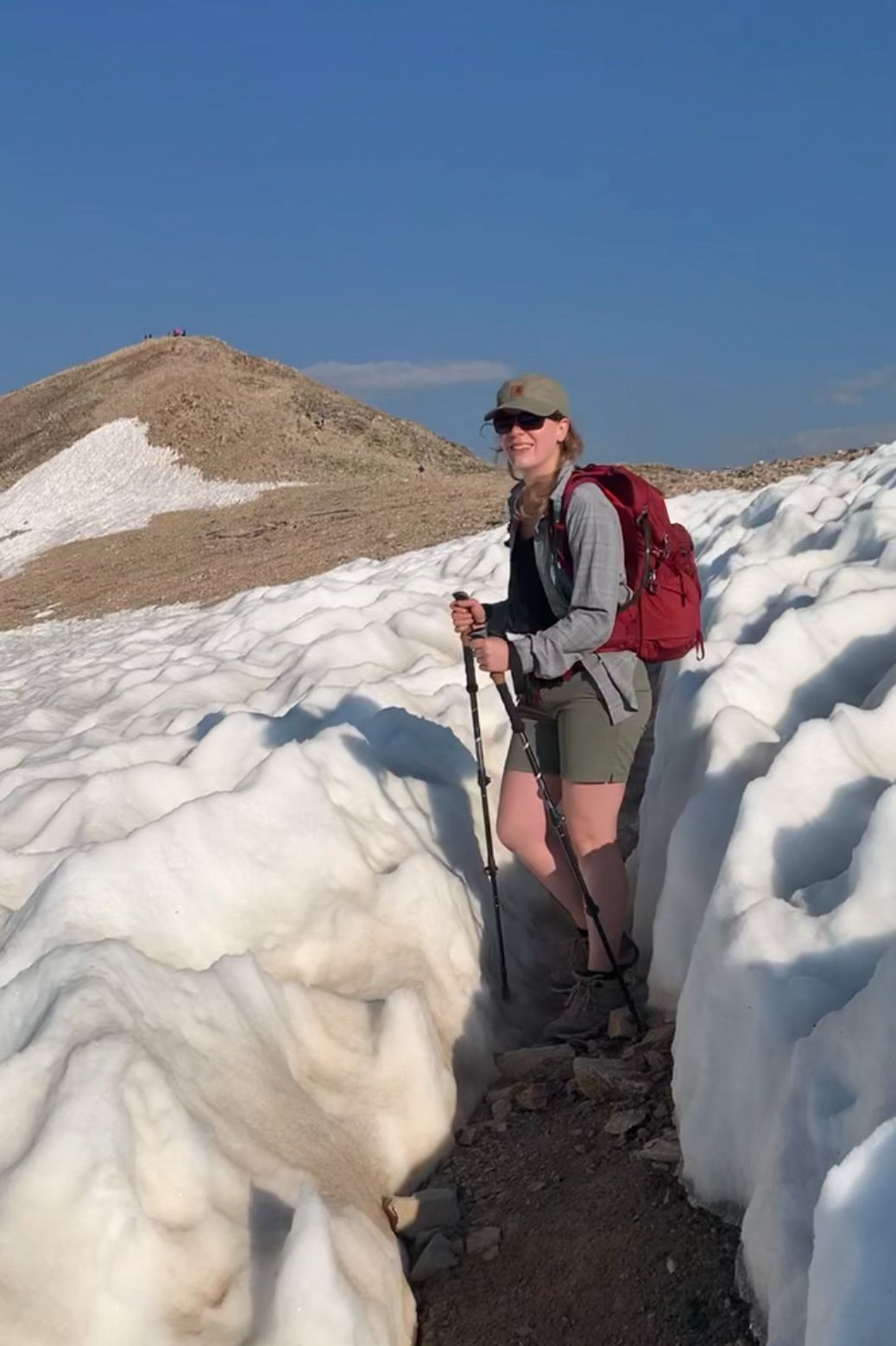 Adventure hiker poses on narrow dirt path between towering snow walls in mountain terrain.