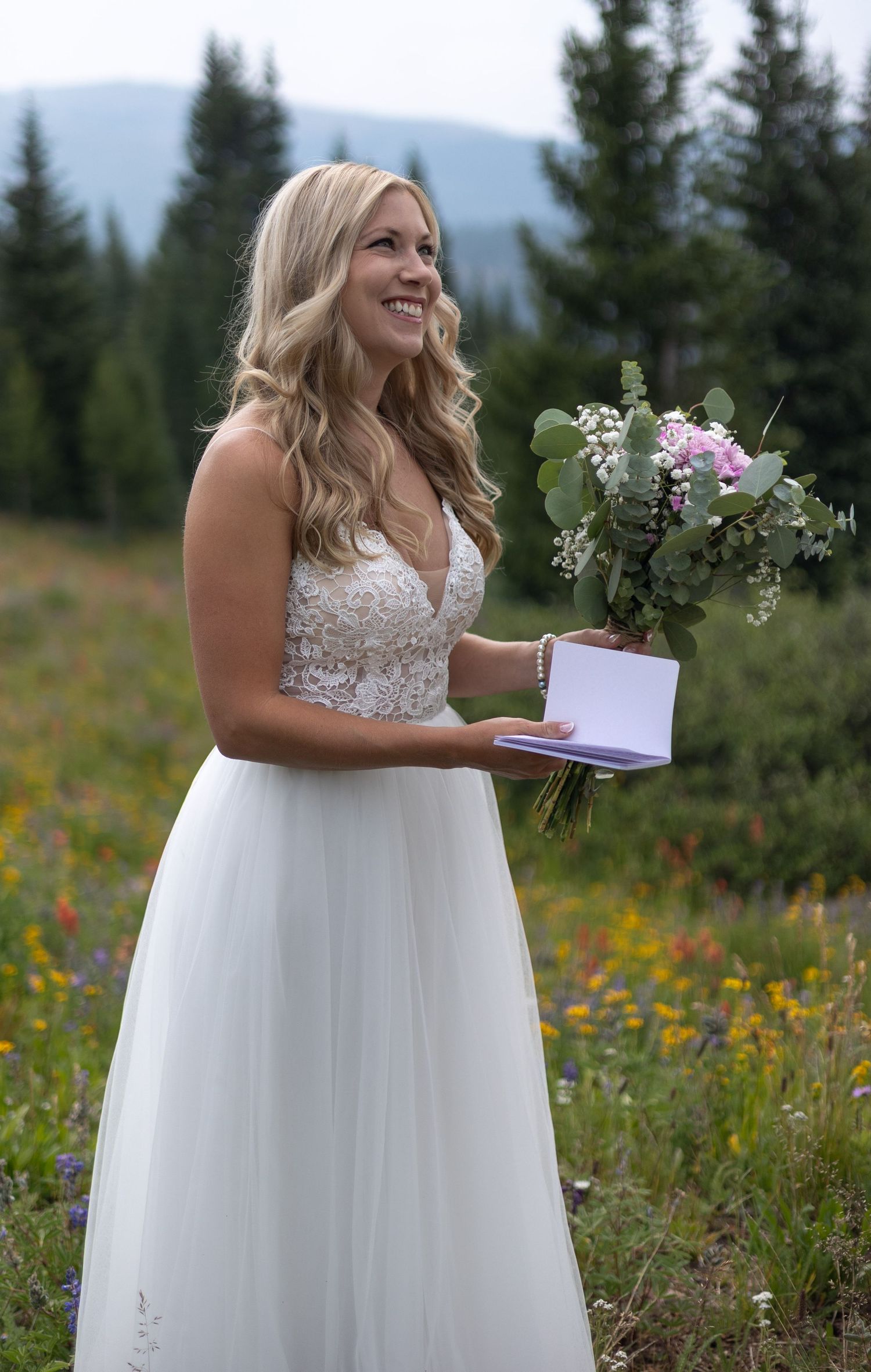 A bride in a white lace gown stands in a wildflower meadow with evergreen trees in the background.