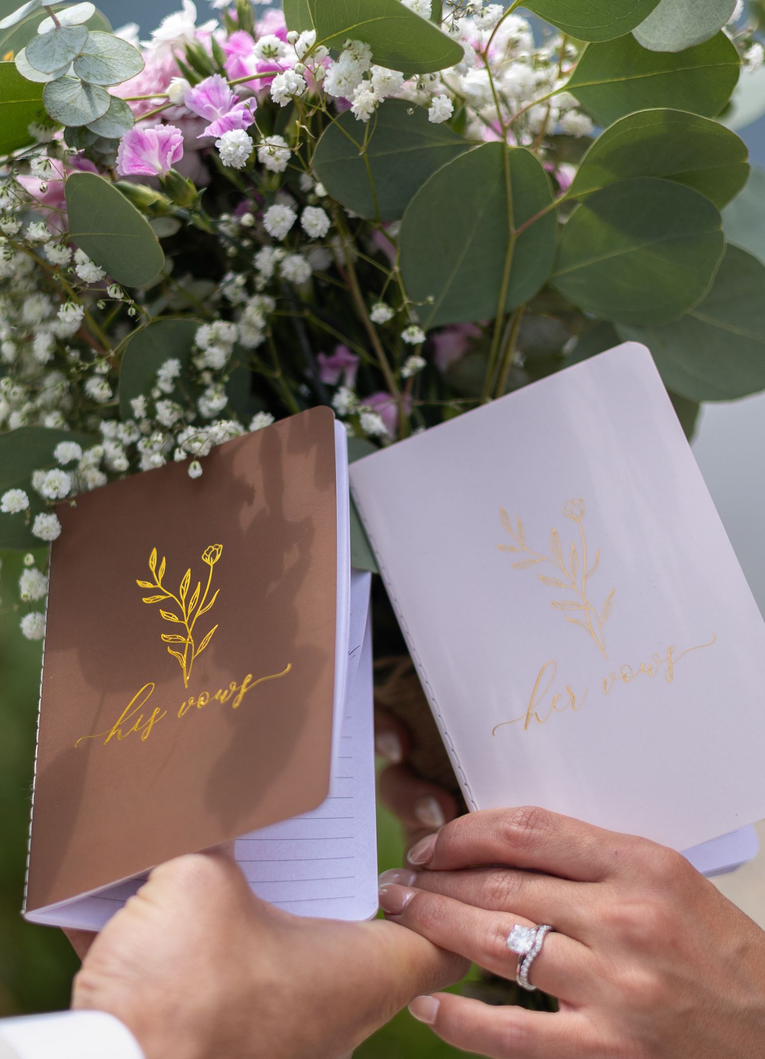 A hand holds a brown and gold wedding vow book against a backdrop of delicate white and pink flowers.