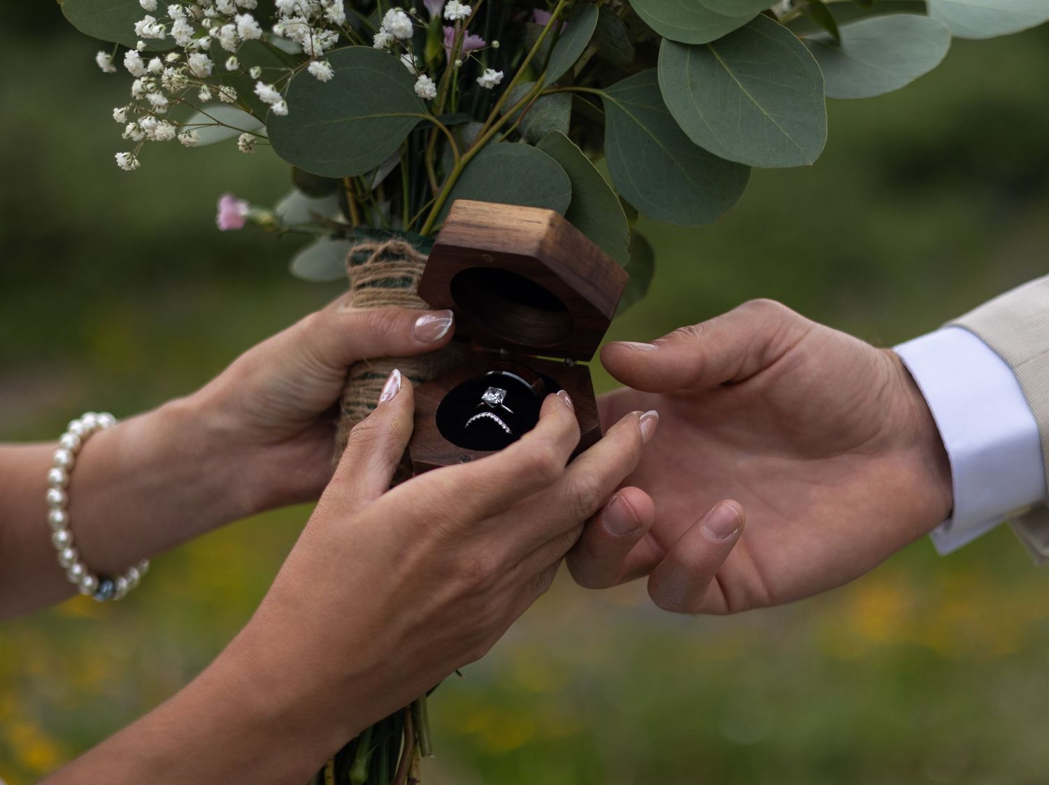 A close-up shot of hands exchanging a wedding ring during an outdoor ceremony with greenery in the background.