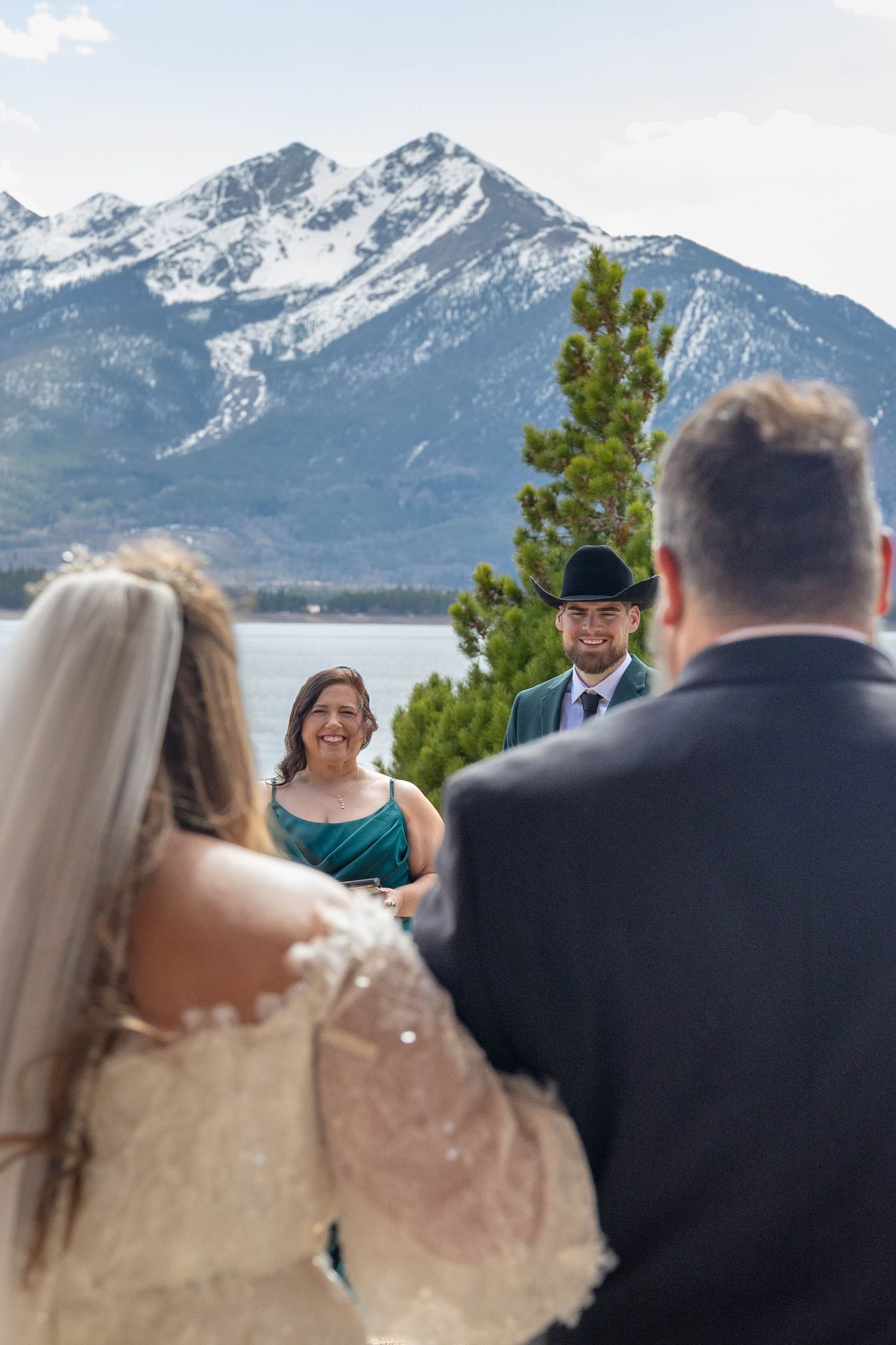 Outdoor mountain wedding ceremony by a lake with snow-capped peaks, groom in black cowboy hat smiling at bride.