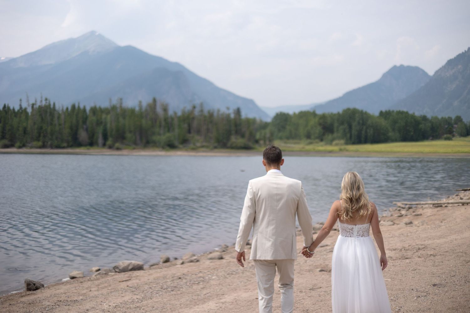 A couple walks hand in hand along a scenic mountain lake shore with evergreen forests and peaks in the background.