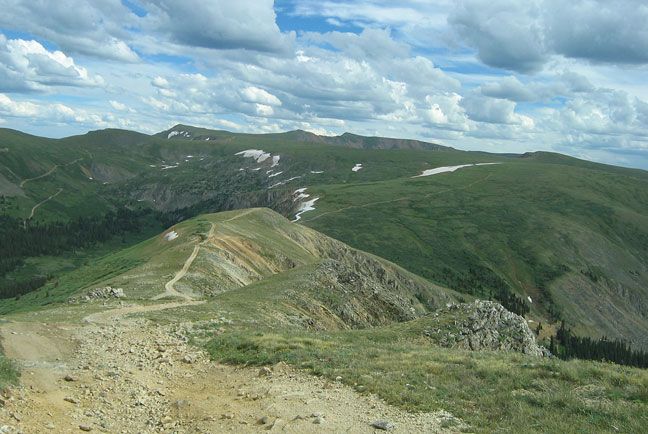 Winding dirt trail along a rocky mountain ridge with green alpine slopes, patches of snow, and a partly cloudy blue sky overhead.