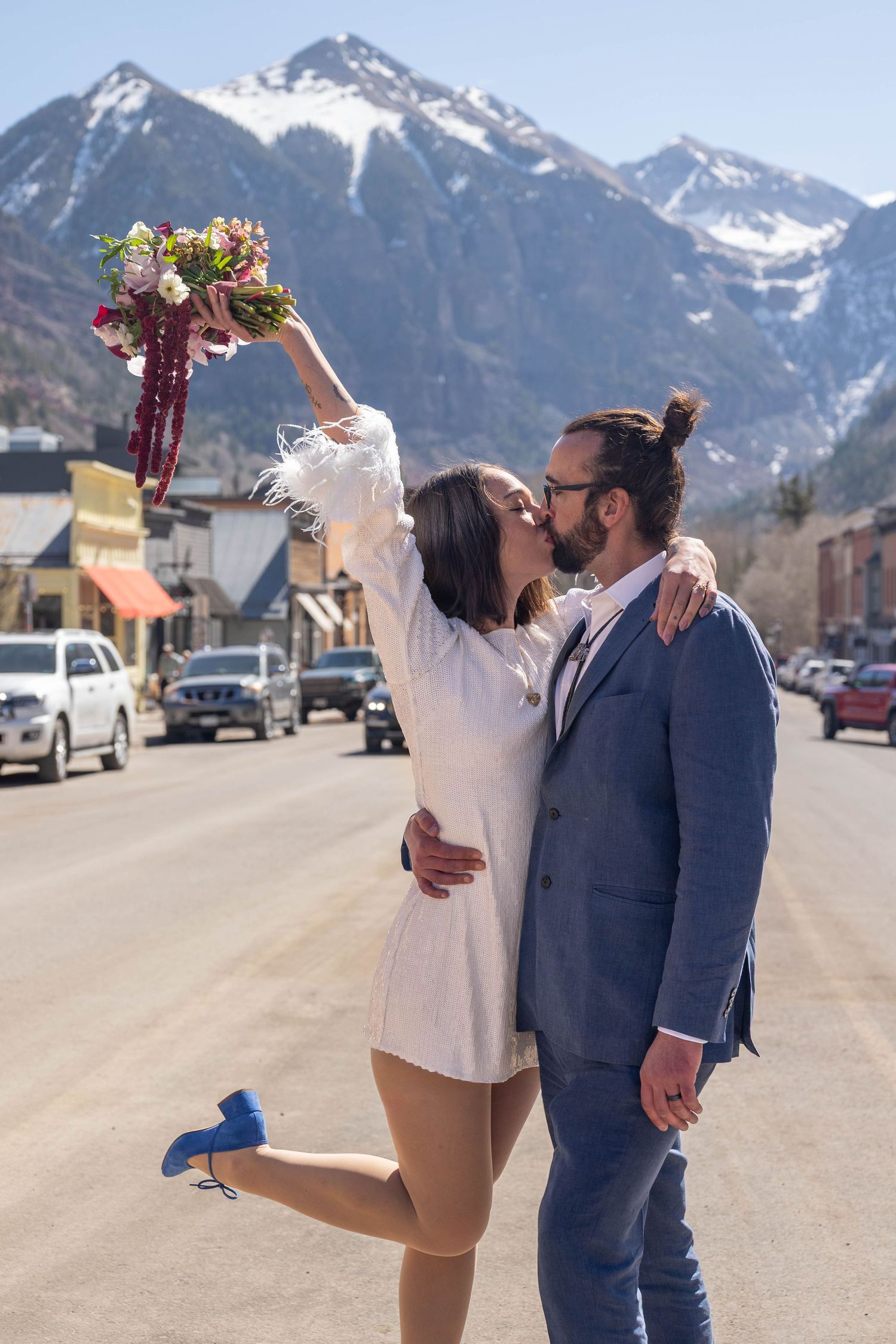 Couple kissing on sunny mountain town street, woman raising floral bouquet while lifting one leg.