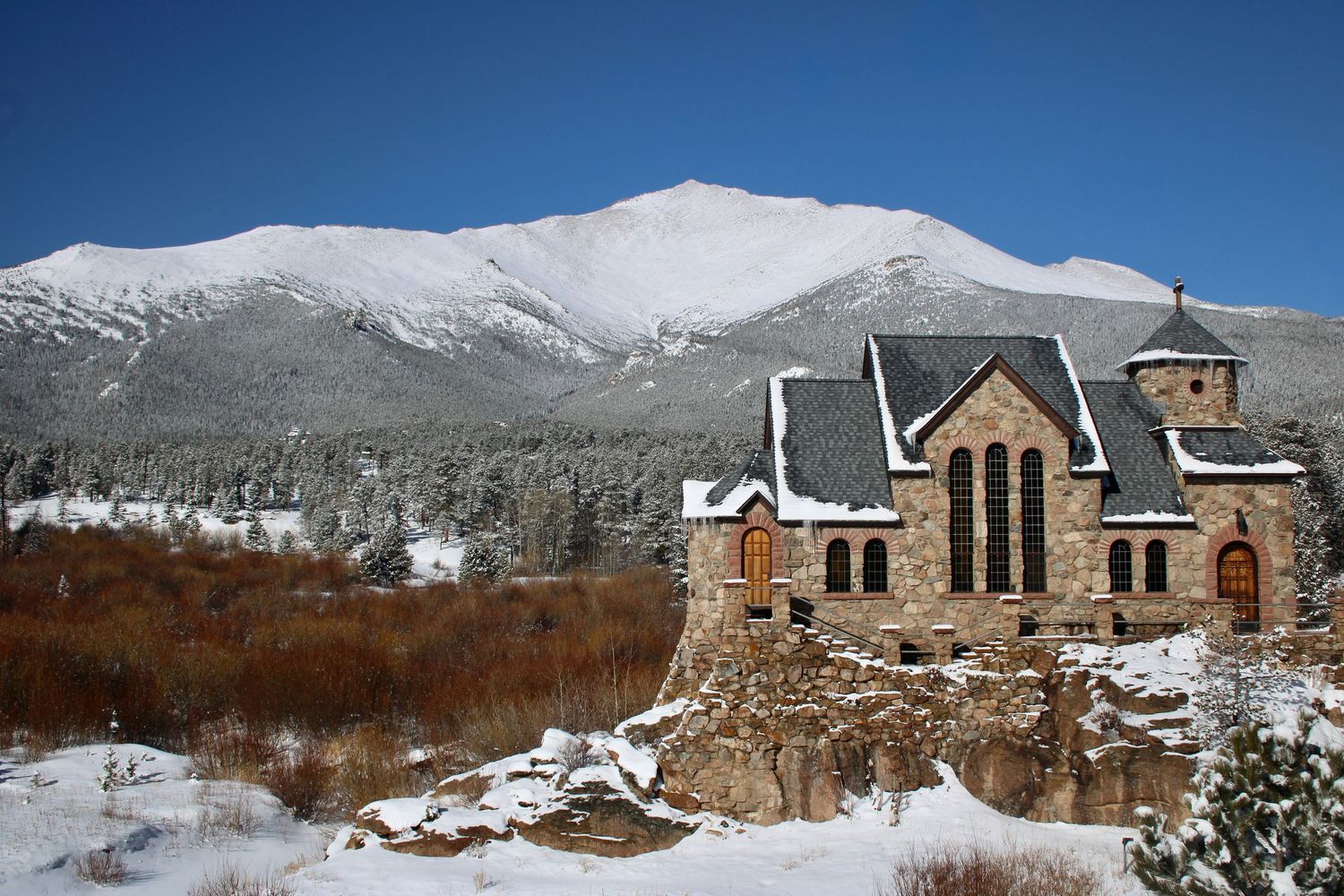 Historic stone chapel with Gothic arched windows sits in snowy Colorado landscape with snow-capped Rocky Mountains behind.