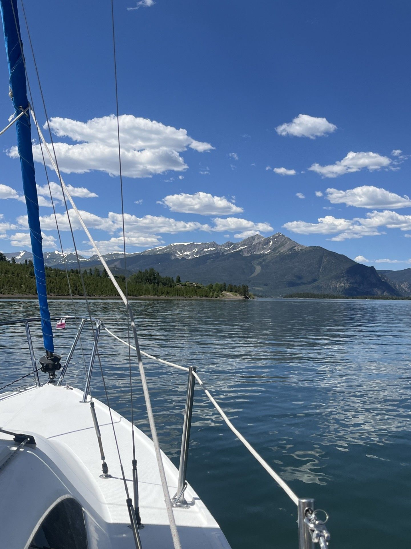 Sailboat bow on calm mountain lake with snow-capped peaks and blue sky with white clouds in background.