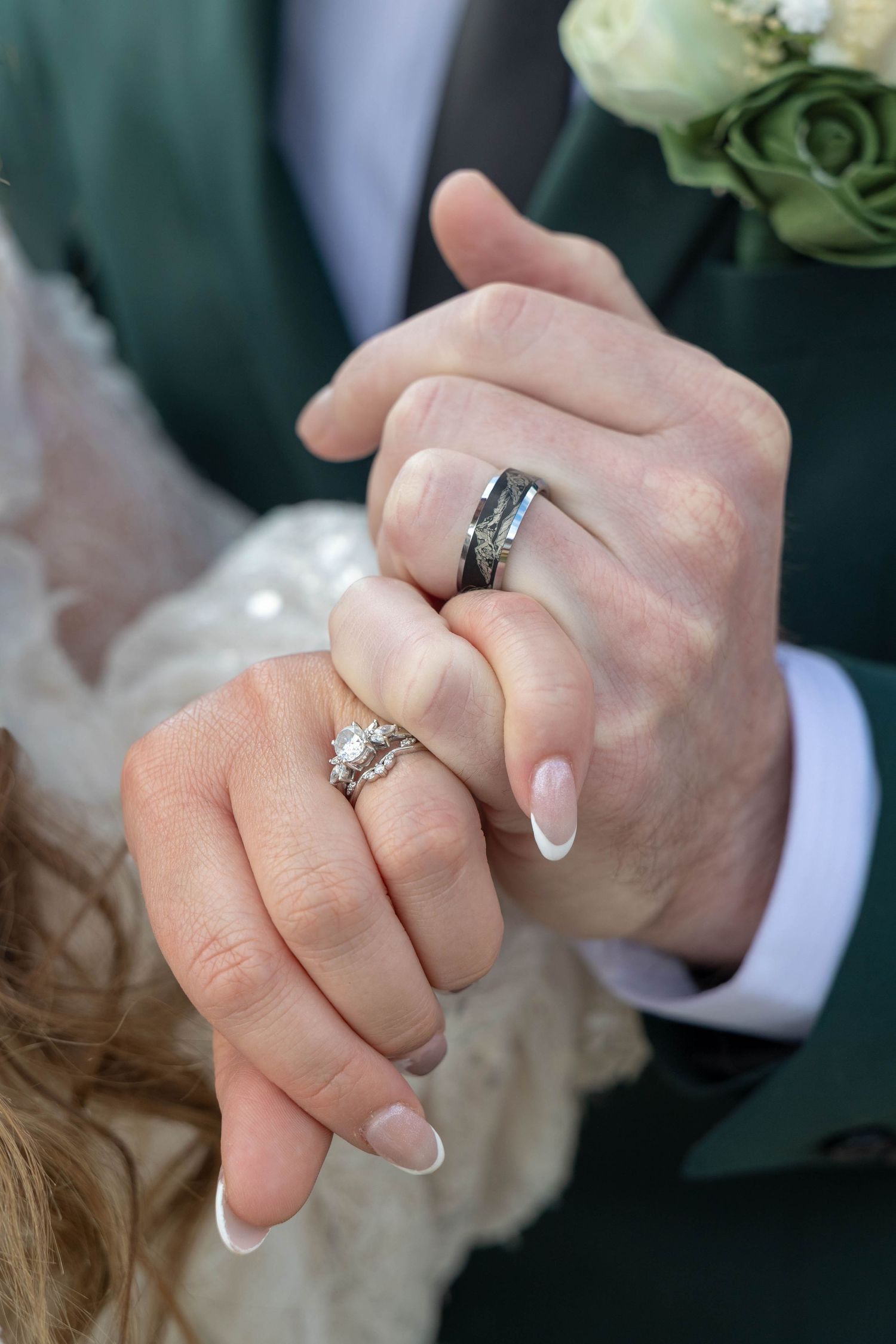 Newlywed couple holding hands showing wedding rings, bride with diamond ring and groom with silver band, wedding attire visible.