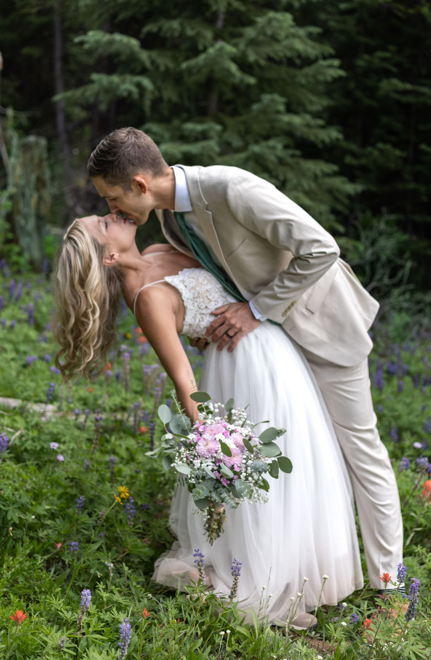 A romantic wedding couple shares an intimate dip kiss in a wildflower meadow surrounded by purple blooms and greenery.