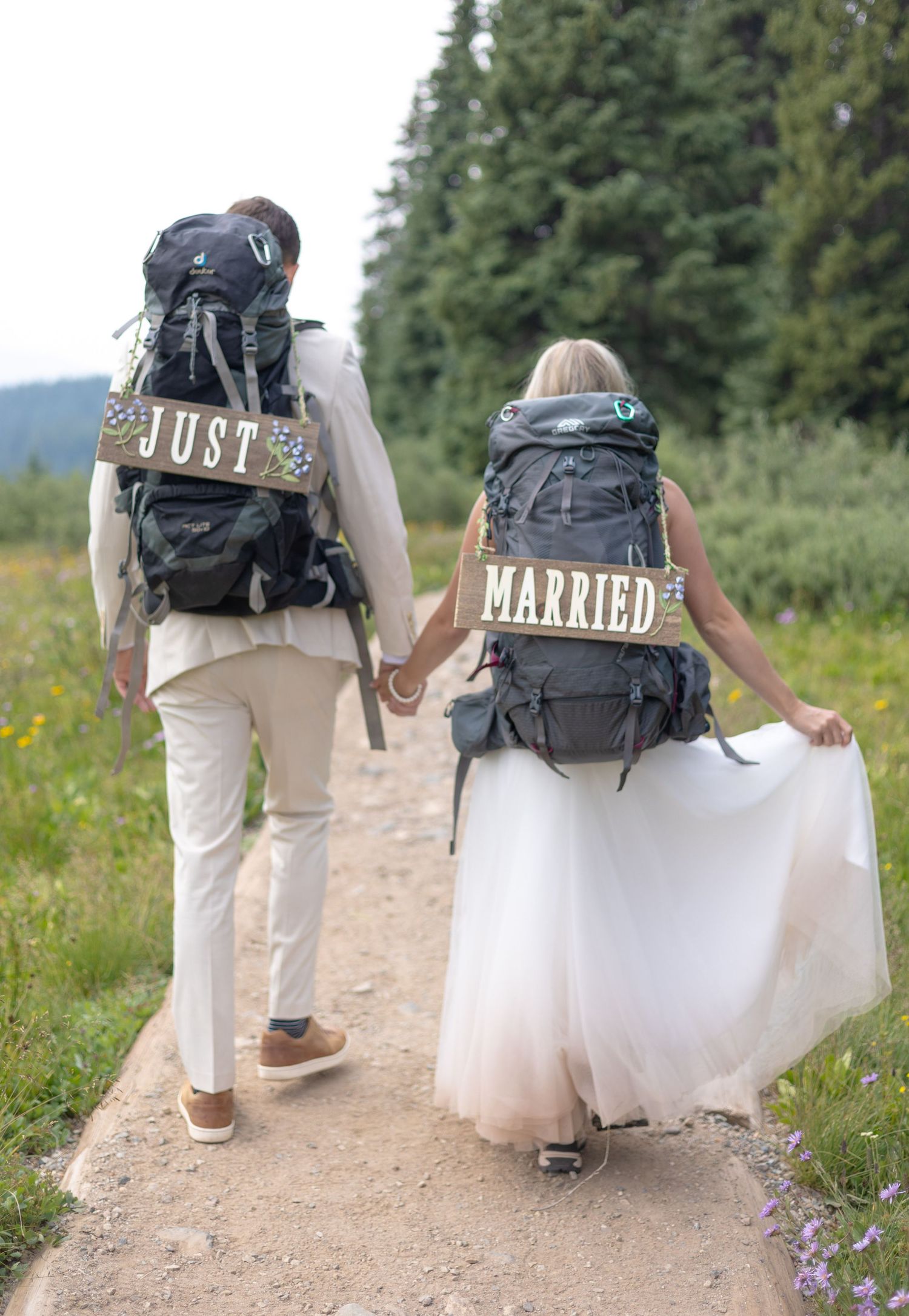A couple wearing backpacks with 'Just Married' text walk together on a mountain trail in wedding attire.