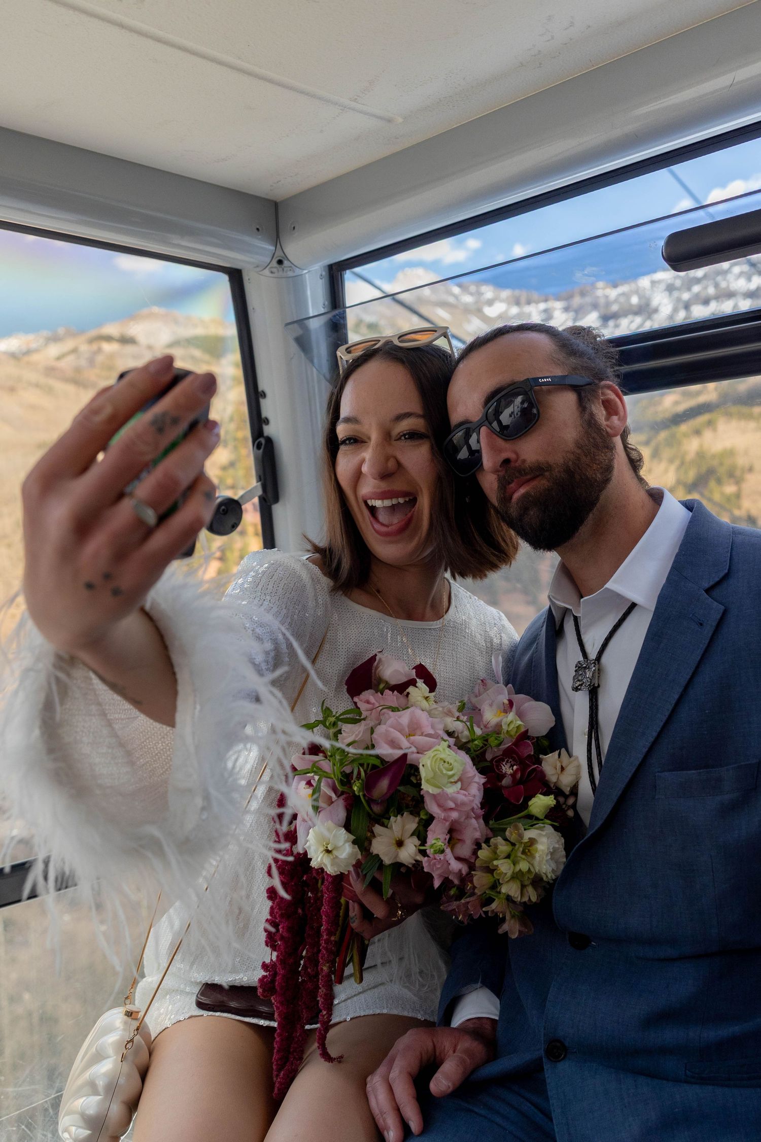 Newlyweds take a selfie in a gondola, bride in white feathered dress holding pink and burgundy bouquet.