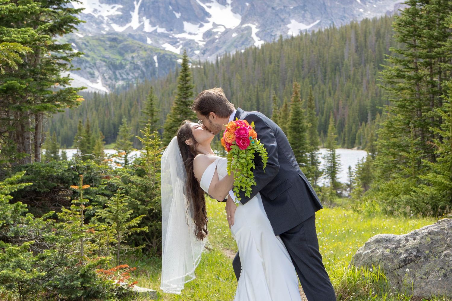 Bride and groom kissing outdoors with mountain backdrop, pine trees, and colorful bouquet during scenic wedding ceremony.