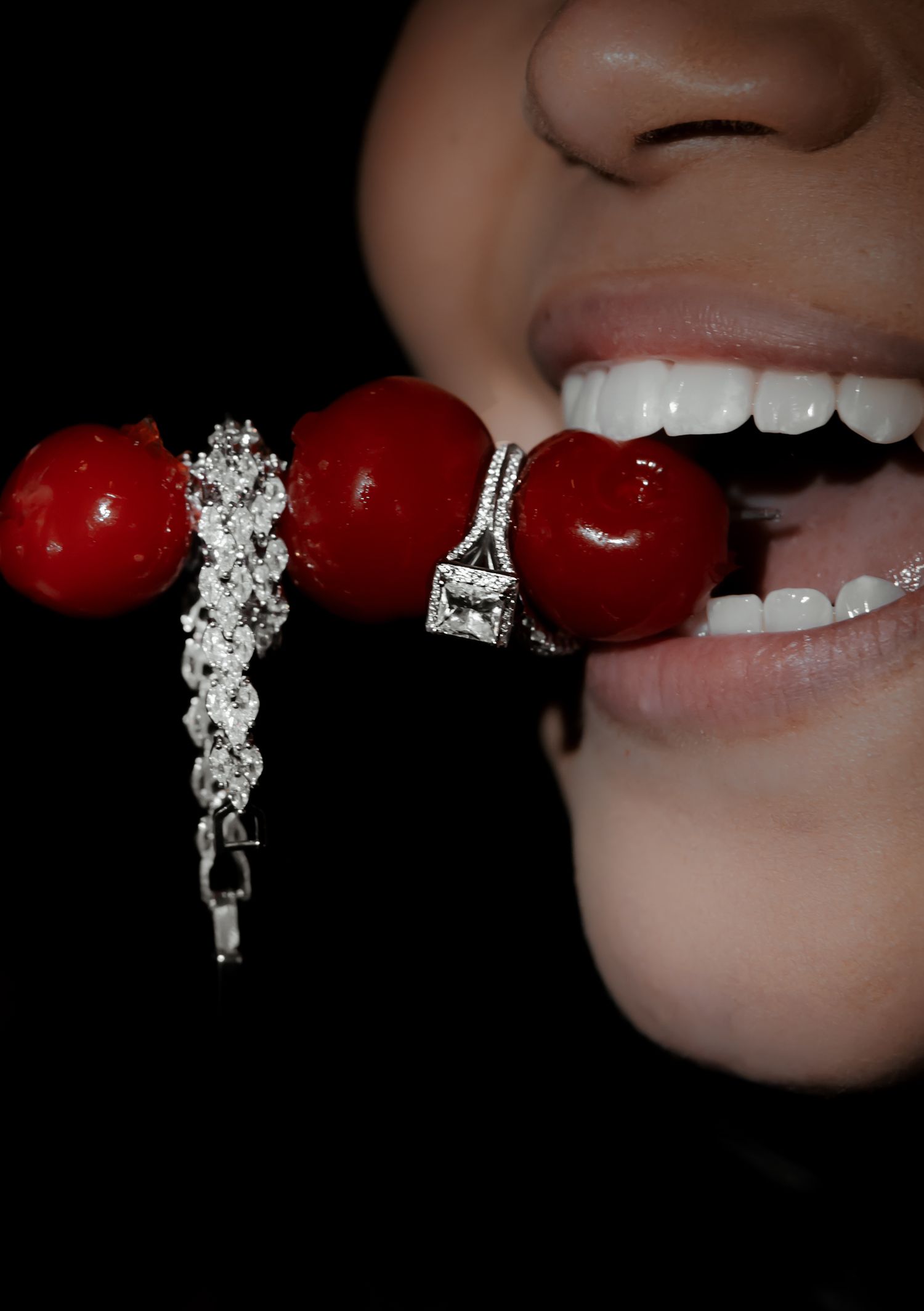 Close-up of a woman biting into red cherries adorned with a sparkling silver diamond bracelet against black background.