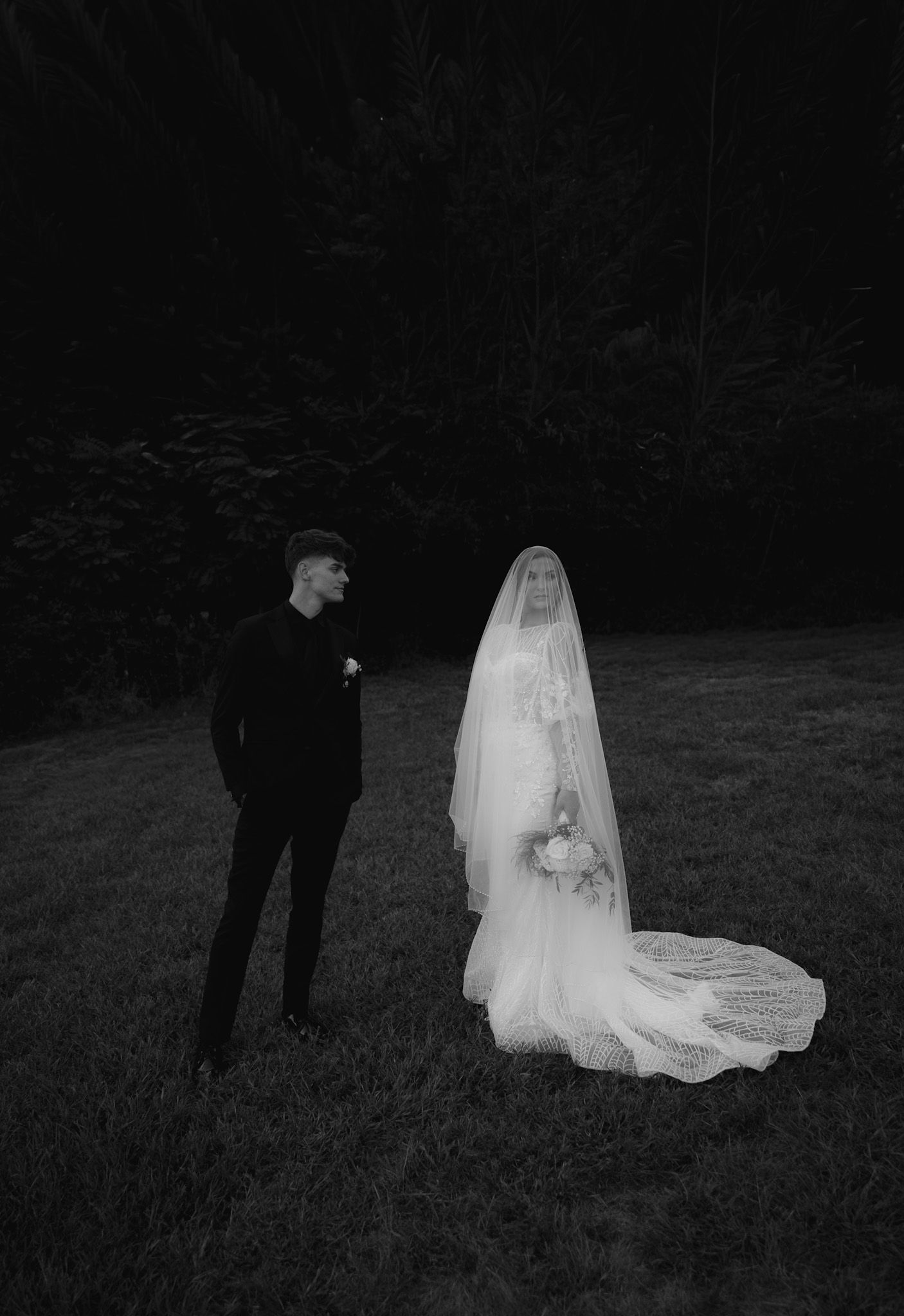 A dramatic black and white photo of a couple in wedding attire standing in a dark grassy field.