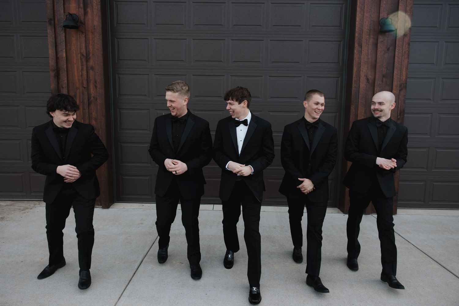 Row of groomsmen in black suits shown from waist down against a dark wall.