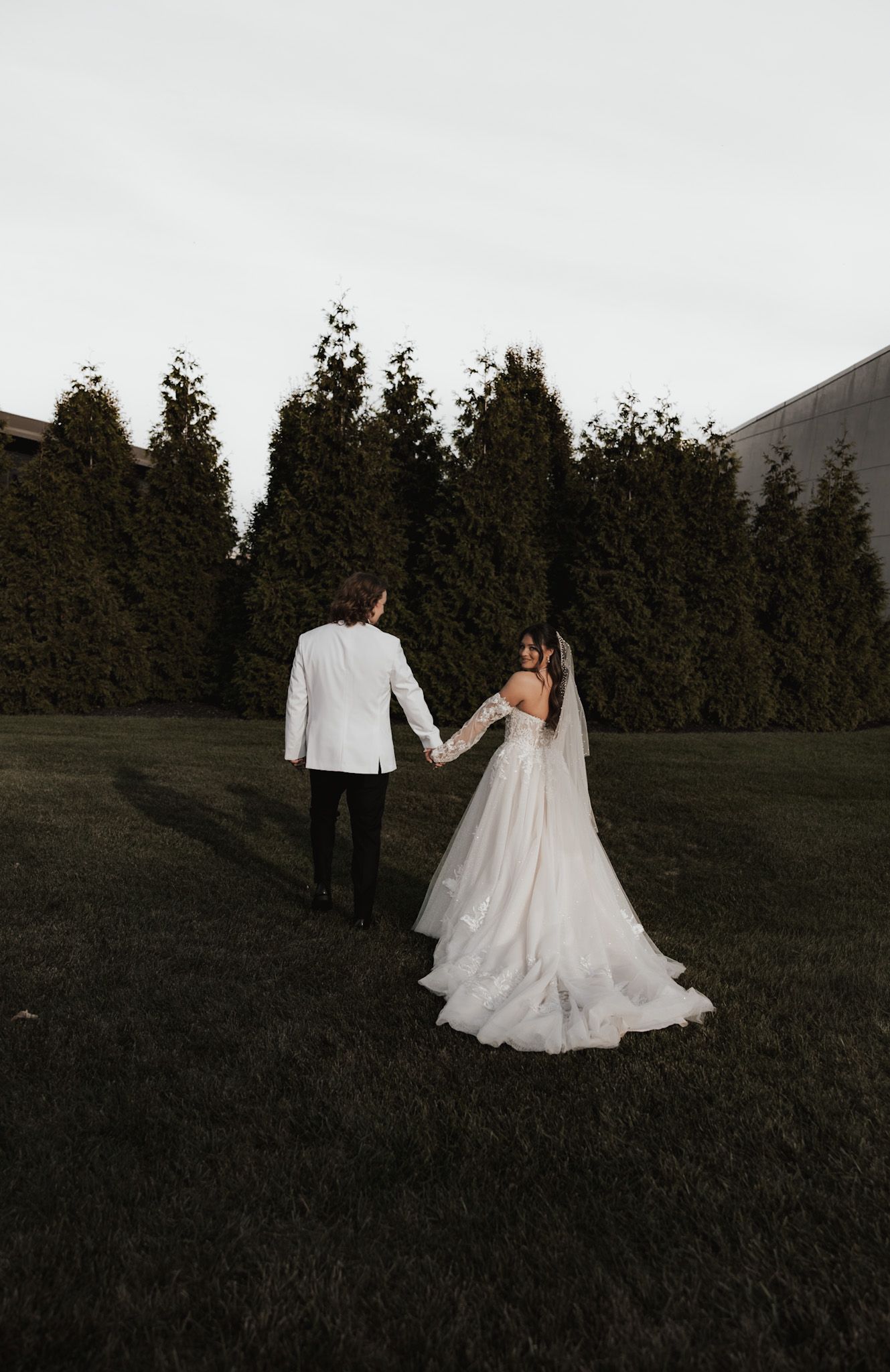 Couple walks through grassy hillside with evergreen trees in moody evening light.