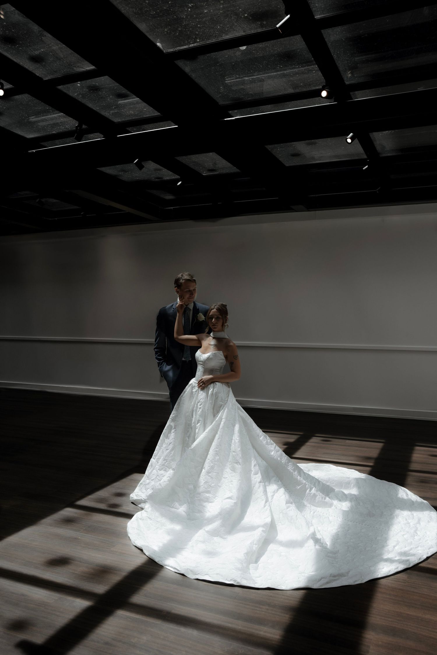 A bride in a flowing white ballgown with a long train stands with a groom in a dark suit in an elegant venue.