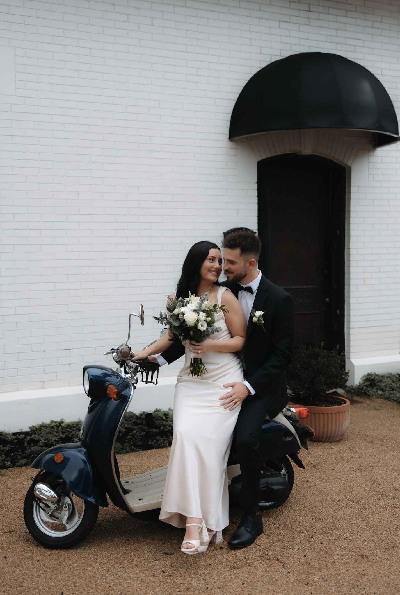 Romantic couple poses with vintage motorcycle against white brick wall.