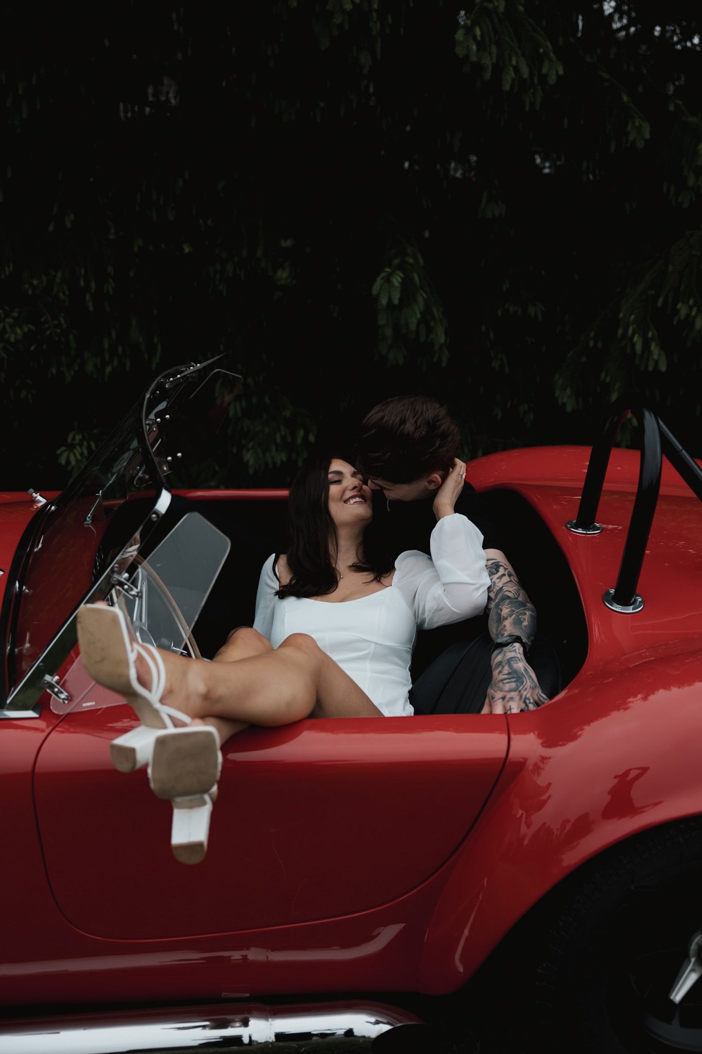 A couple lounging in a vintage red sports car under tree cover.