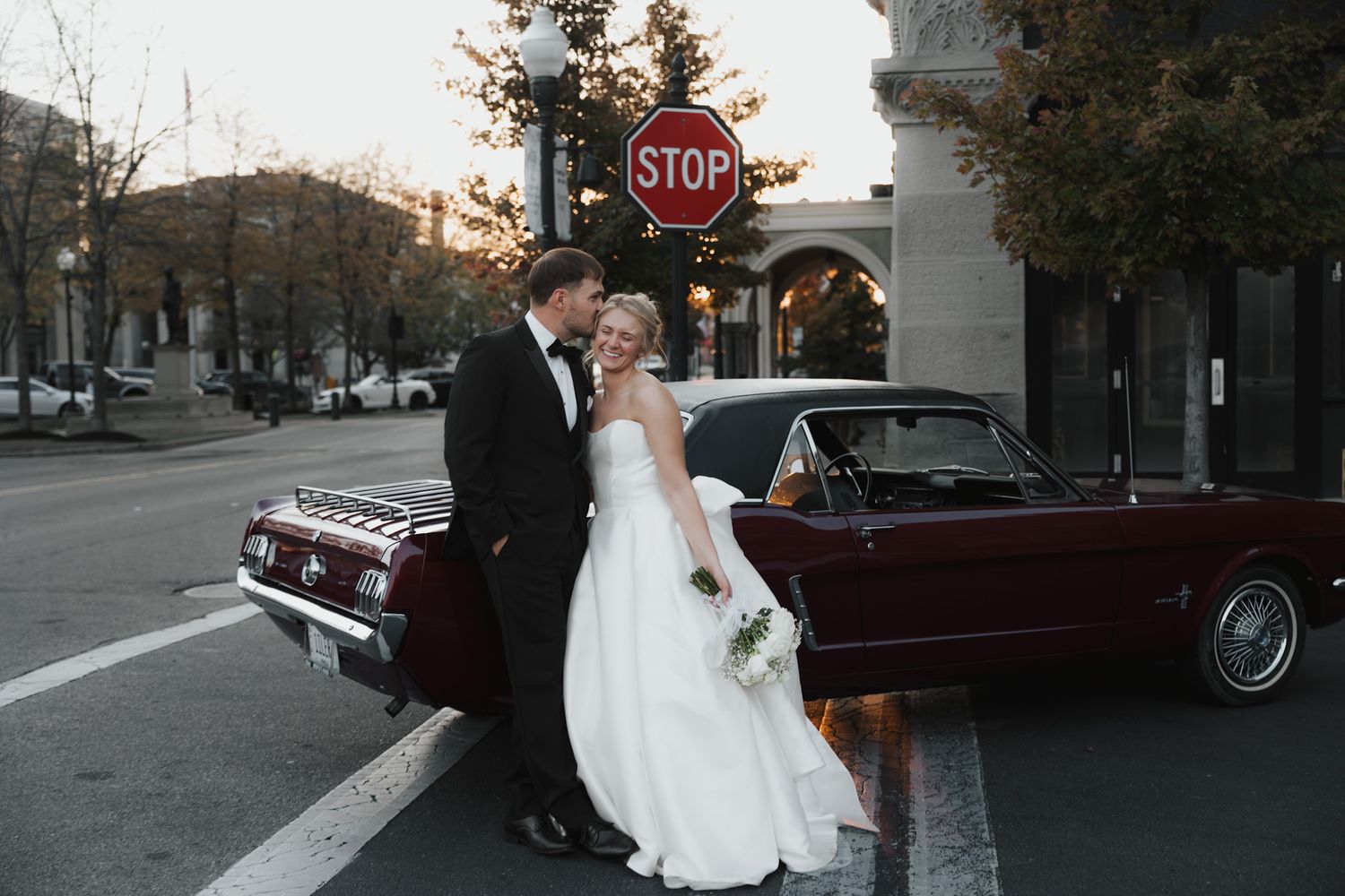 Wedding couple posing next to vintage car on city street with stop sign.