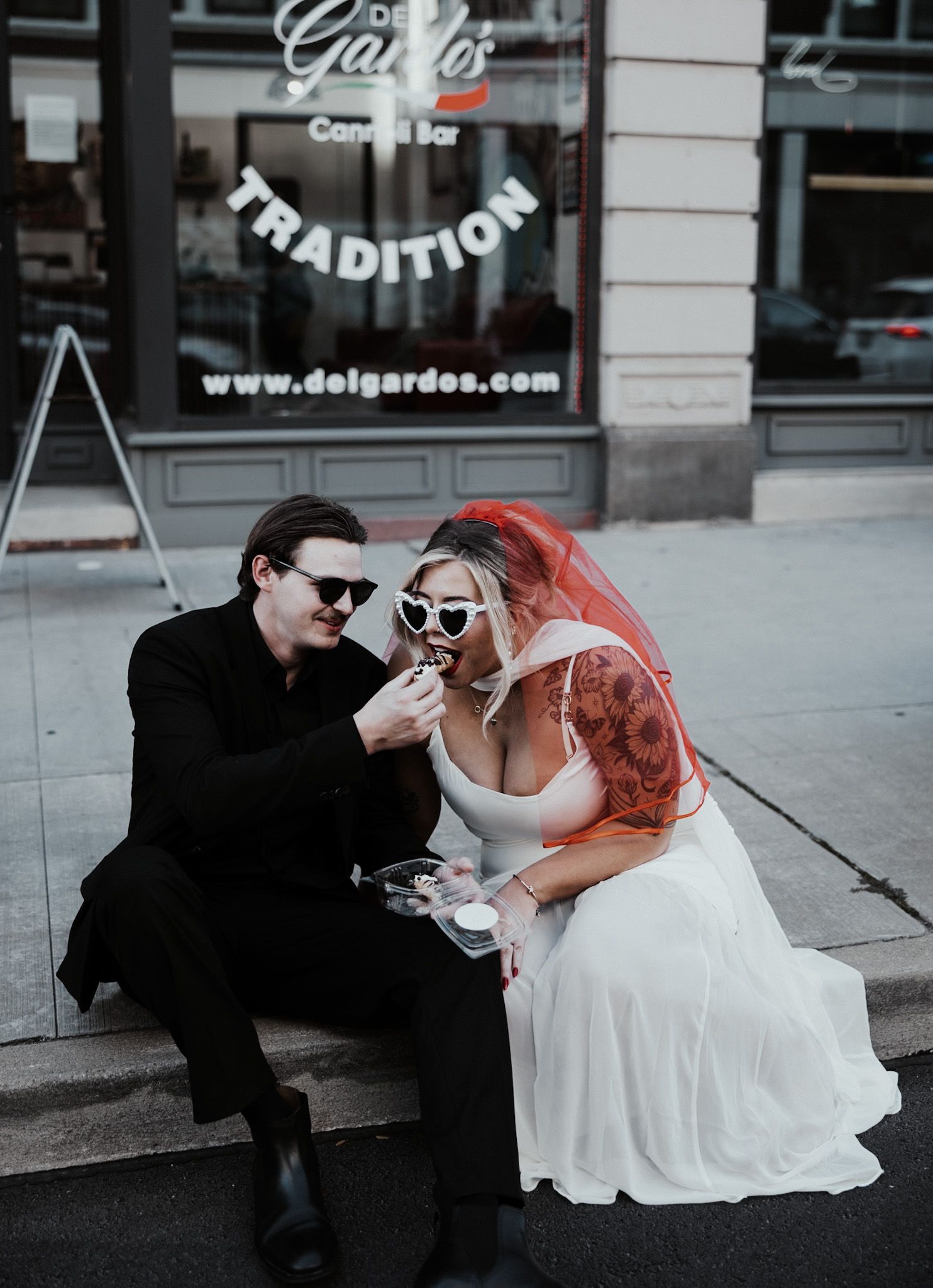Romantic couple in wedding attire sharing a drink outside a storefront.
