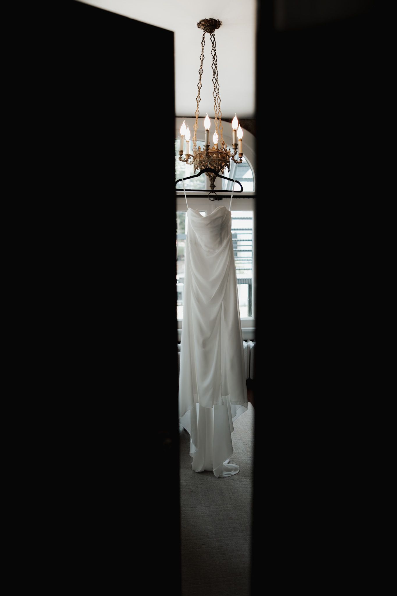 Wedding dress hanging beneath crystal chandelier, viewed through doorway.
