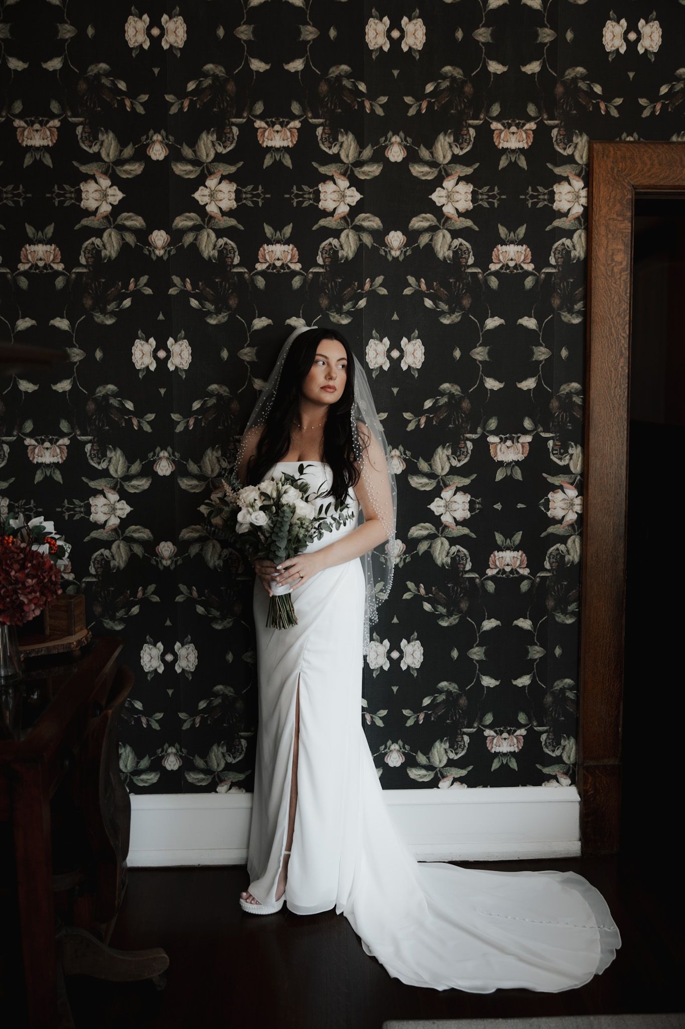 A bride in a flowing white gown poses against a floral wallpaper background.