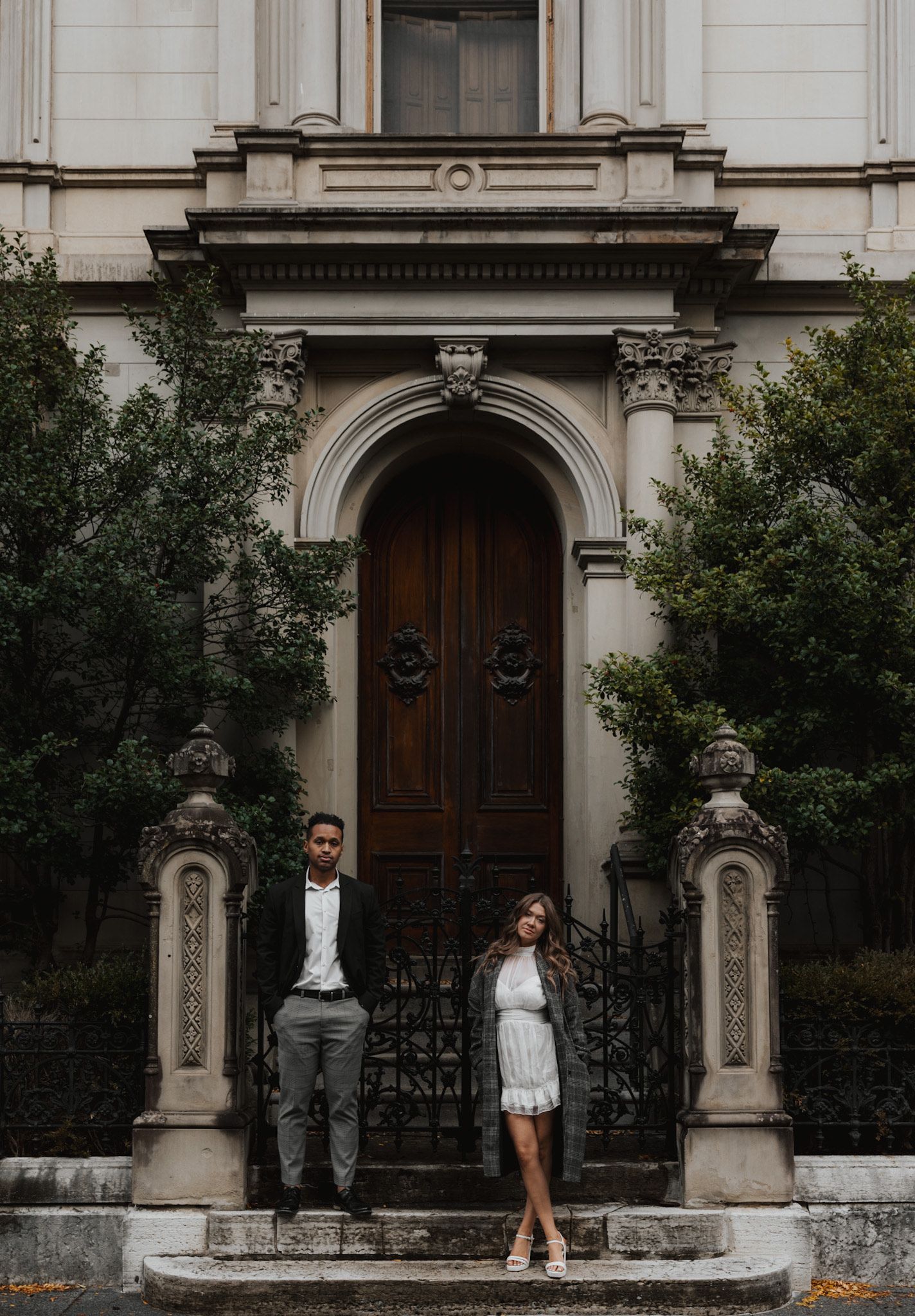 Stylish couple posing in front of ornate historic doorway with stone archway and steps.