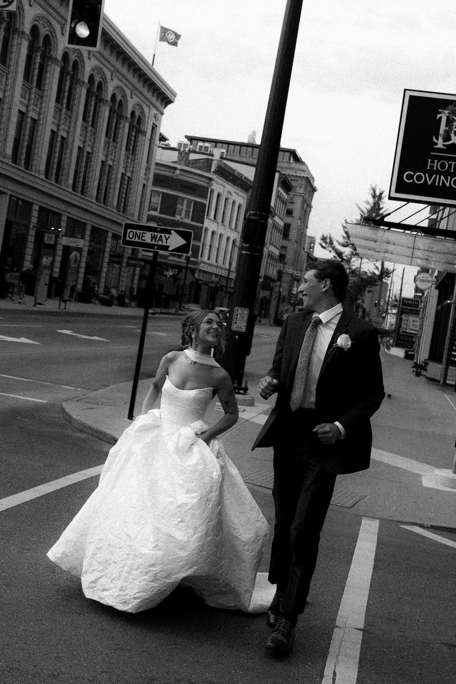 Black and white photo of a bride and groom crossing a city street corner, laughing together on their wedding day.