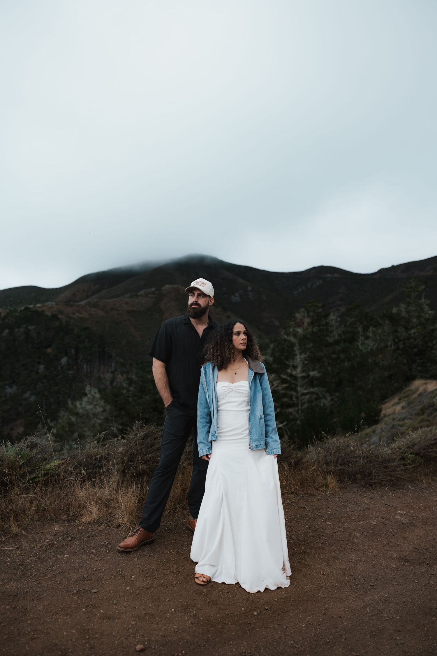 Casual couple standing on mountain trail with foggy peaks in background.
