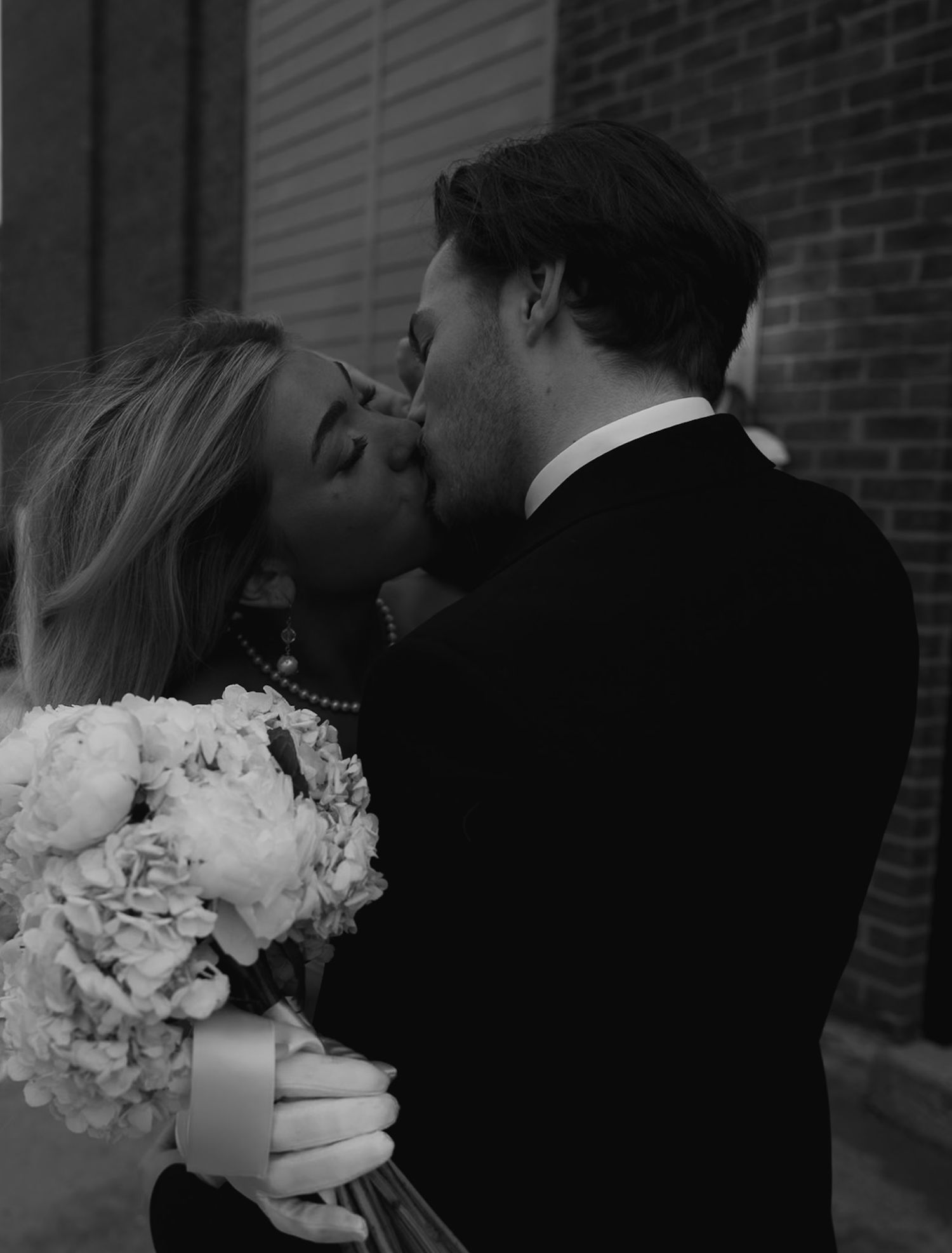 Couple sharing intimate moment with white floral bouquet against brick wall.