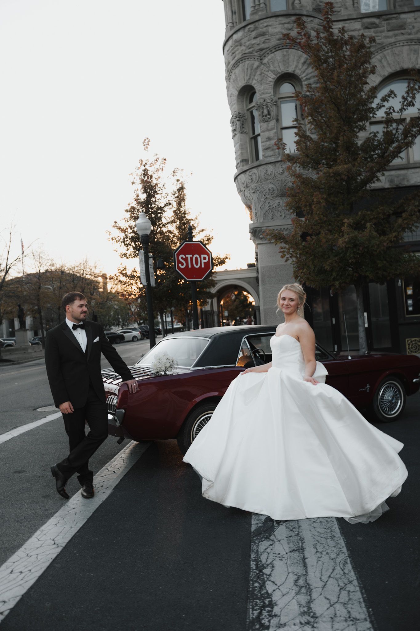 A bride in a flowing white dress poses near a vintage car on a city street.
