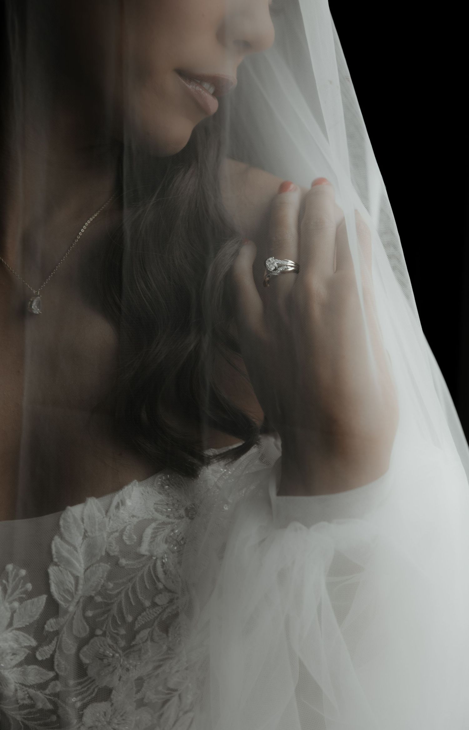 Bride in lace wedding dress and veil, showcasing a diamond engagement ring while holding her veil close.