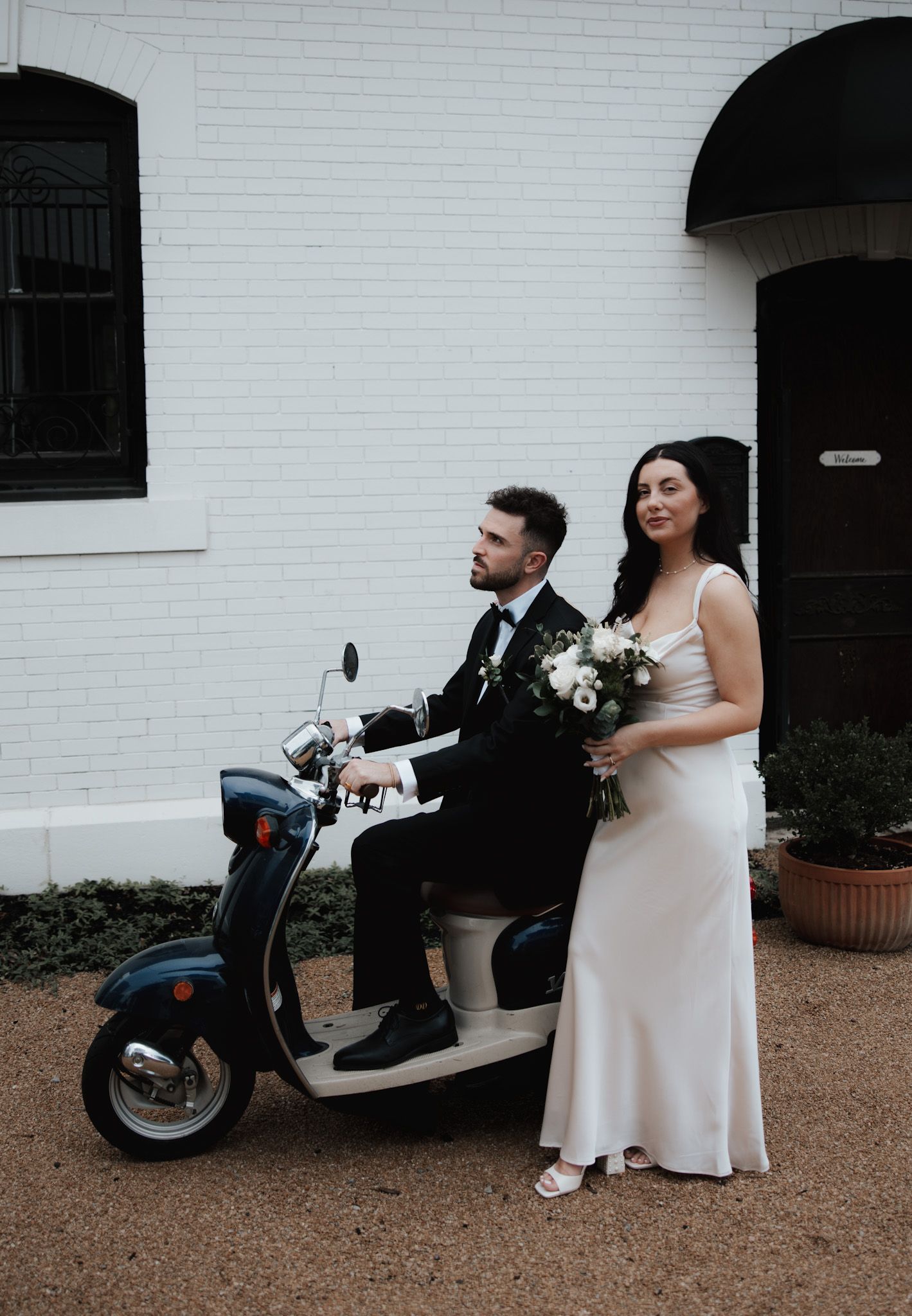 A couple poses with a white vintage scooter against a white brick wall.